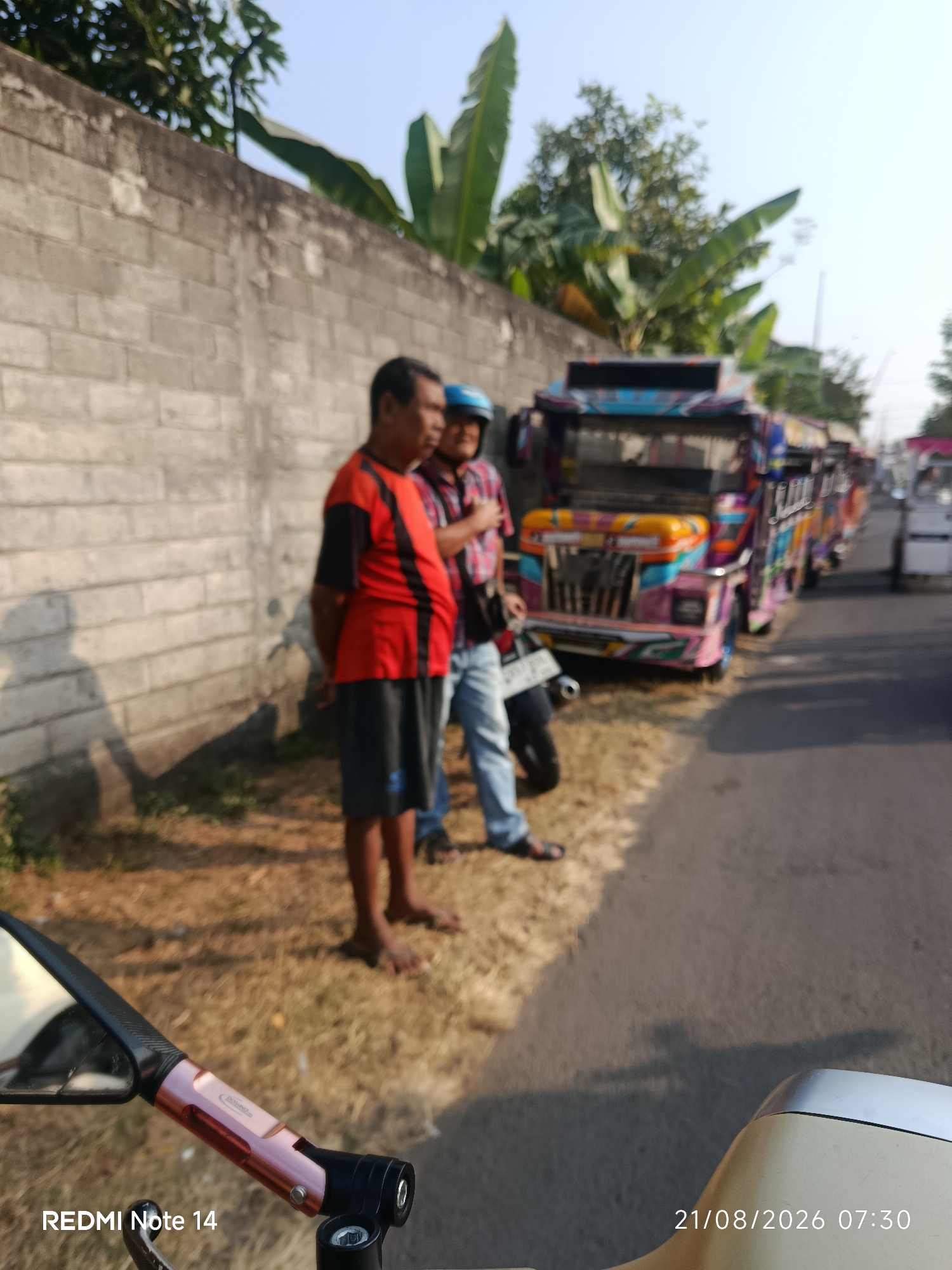 The image shows a street scene in Panggungrejo, Indonesia, with a row of colorful, decorated vehicles, possibly minibuses or modified jeeps, parked along the side of the road. Two men are standing near a wall to the left of the frame. One man is wearing a red and black striped shirt and dark shorts, while the other is wearing a plaid shirt and blue jeans and has a blue helmet on. The vehicles appear to be decorated with bright colors and intricate patterns. The environment includes a concrete wall, dry grass, and a paved road. A portion of a scooter is visible in the foreground on the left, with a mirror and handlebars. A timestamp "21/08/2026 07:30" and "REDMI Note 14" are present in the bottom left corner. The lighting suggests it is daytime, possibly morning.