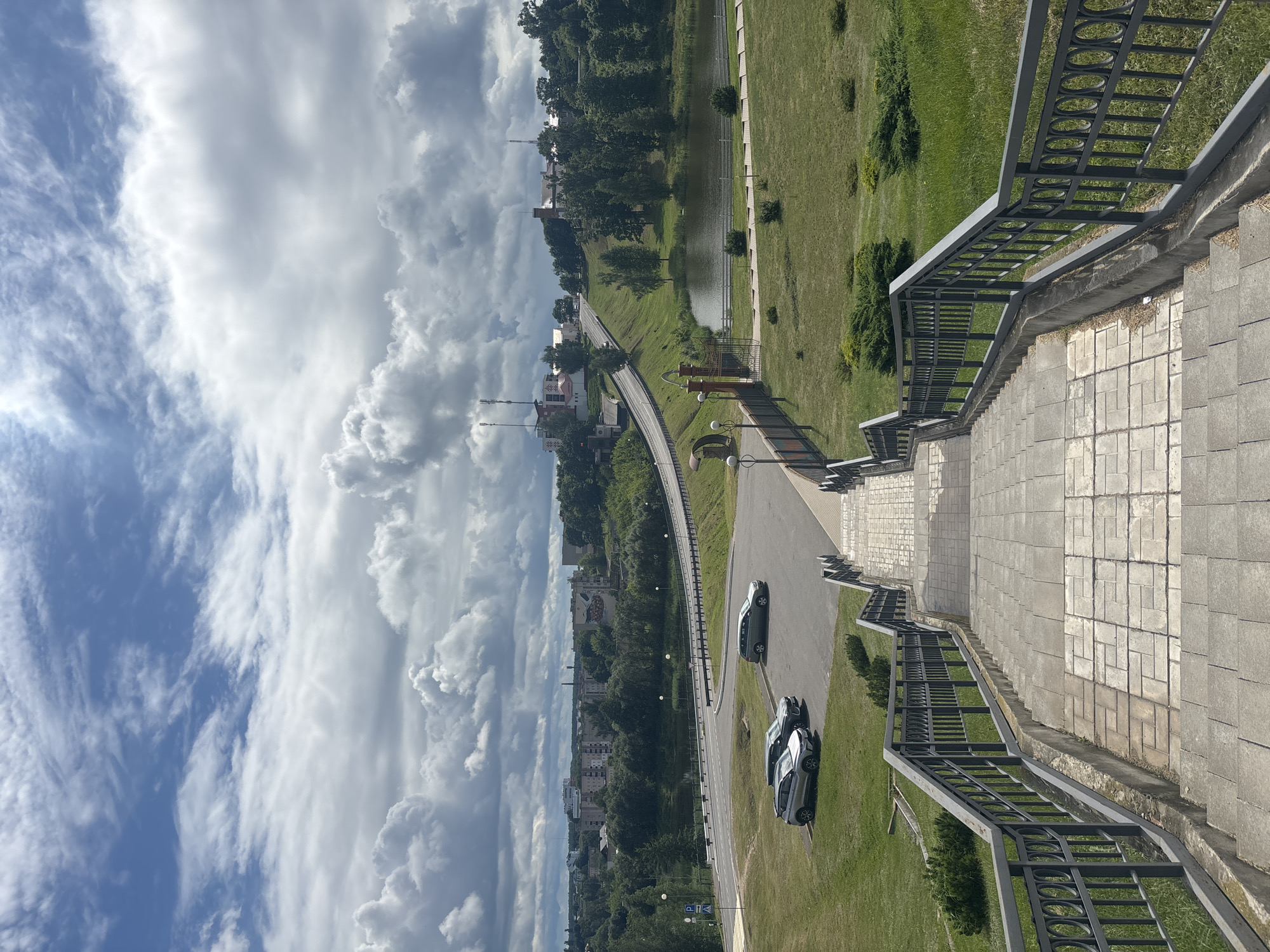 The image shows a wide stone staircase with a dark metal railing leading down from a viewpoint. The staircase is paved with rectangular stone tiles. To the left and right of the staircase, there is a grassy slope with scattered bushes. In the middle distance, there is a road that curves to the right, with two cars parked on a paved area next to the road. Beyond the road, there is a body of water, likely a pond or small lake, with a paved walkway alongside it. Further in the background, there is a wooded area with a town or city visible on a hill. The town features several buildings, including some with red roofs and towers, and what appear to be cell phone towers. The sky is filled with dramatic, dark clouds, suggesting overcast or approaching weather. The overall scene depicts an outdoor public space in a semi-urban or suburban environment. The location is identified as Капыльскі сельскі Савет, Belarus.