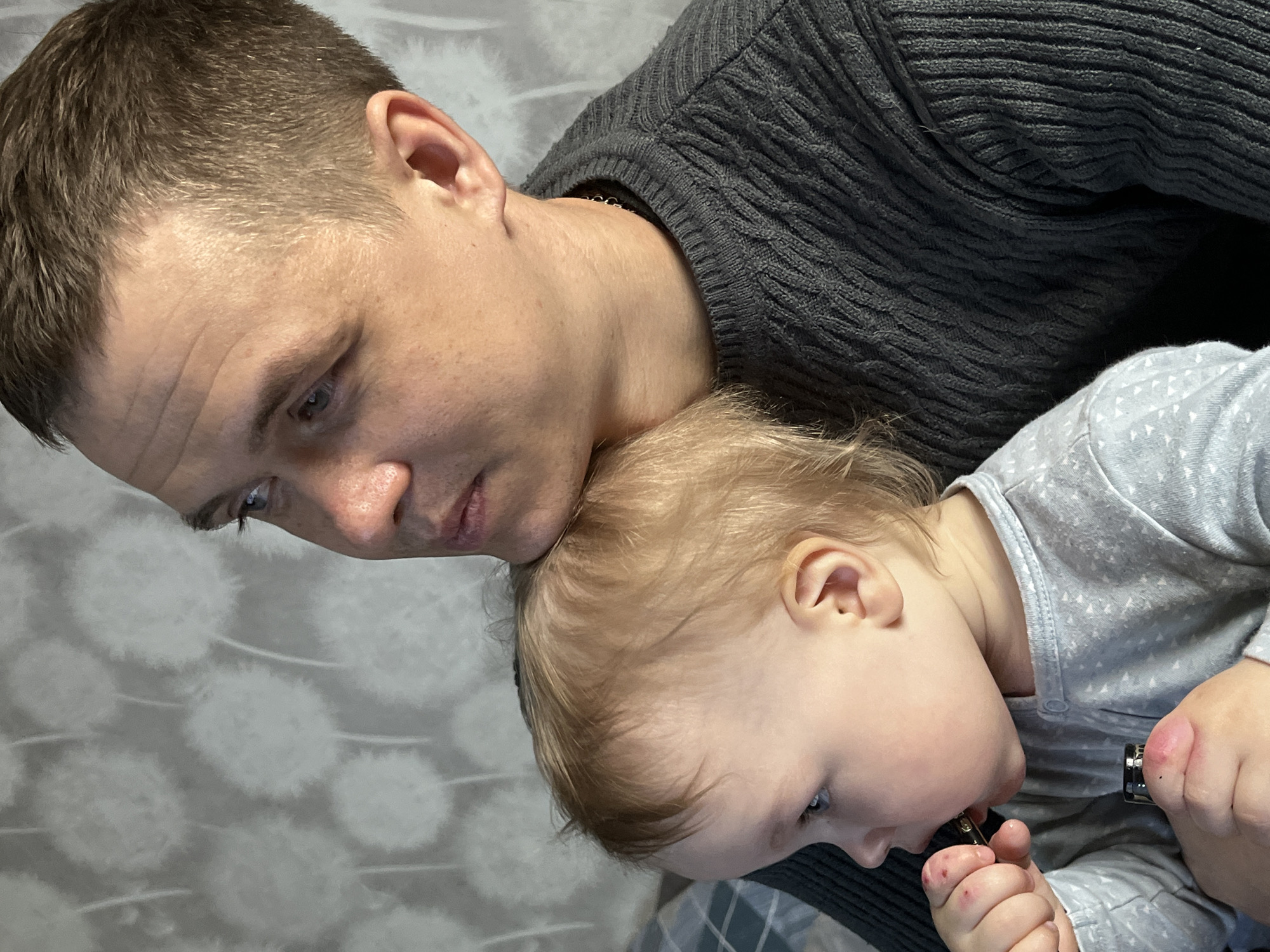 The image shows an adult male and a young child, likely in an indoor setting. The male is fair-skinned with short, light brown hair, wearing a dark gray, textured sweater. He is looking down. The child, also fair-skinned with light blonde hair, rests their head on the man's shoulder. The child is wearing a light gray patterned onesie and is holding a small, dark object in their hand. The background appears to be a patterned wall or fabric. There is no information in the image to establish the location. It is not possible to confirm the location as Chaikovsky, Russia, based on the provided image.
