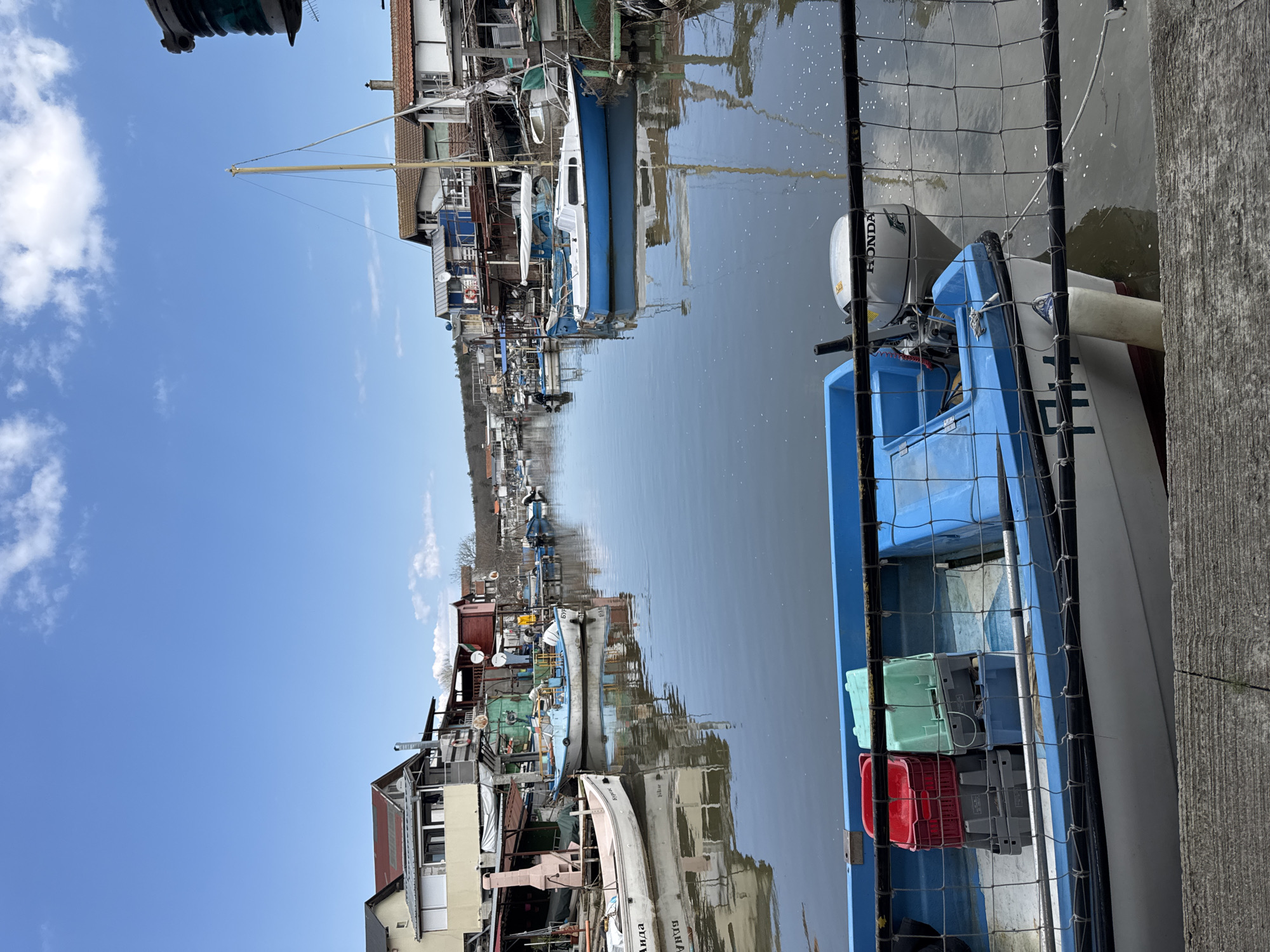 A canal in Burgas, Bulgaria, is filled with boats moored along the sides. Houses, some of them waterfront properties, are visible on both sides of the canal. The sky is clear with some clouds. The canal reflects the sky and the buildings. In the foreground, a blue and white boat is docked, with plastic crates stacked inside. A metal fence separates the boat from the canal.