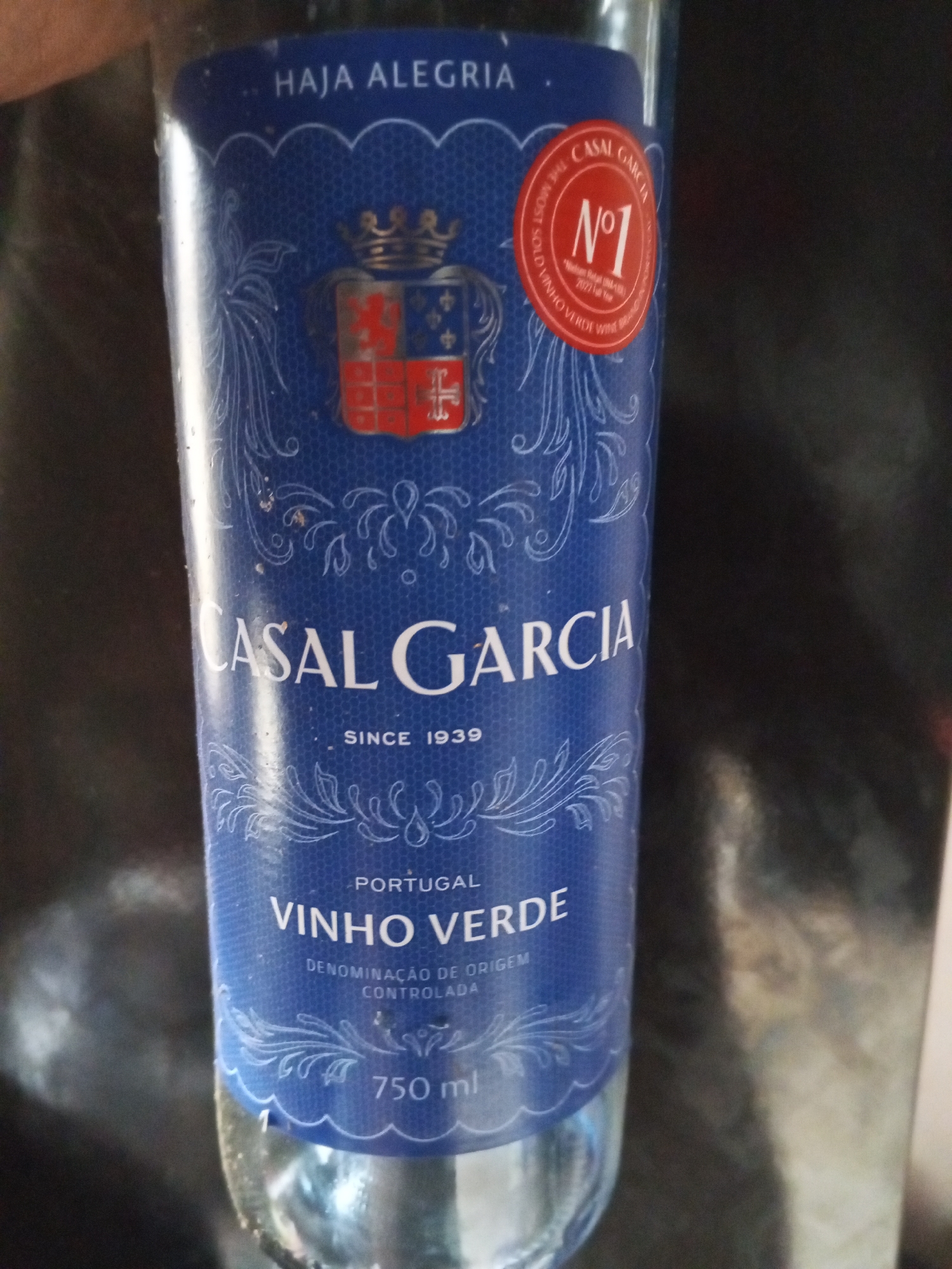 A close-up shot shows a blue label on a glass bottle of wine. The label features a crest with a crown, a red and blue shield, and decorative scrollwork. Text on the label reads "HAJA ALEGRIA," "CASAL GARCIA," "SINCE 1939," "PORTUGAL," "VINHO VERDE," and "DENOMINAÇÃO DE ORIGEM CONTROLADA." A circular red sticker on the upper right of the label states "CASAL GARCIA N°1 THE MOST SOLD VINHO VERDE WINE." The bottle is partially filled with a clear liquid and is standing upright against a dark, reflective background. The environment appears to be indoors.