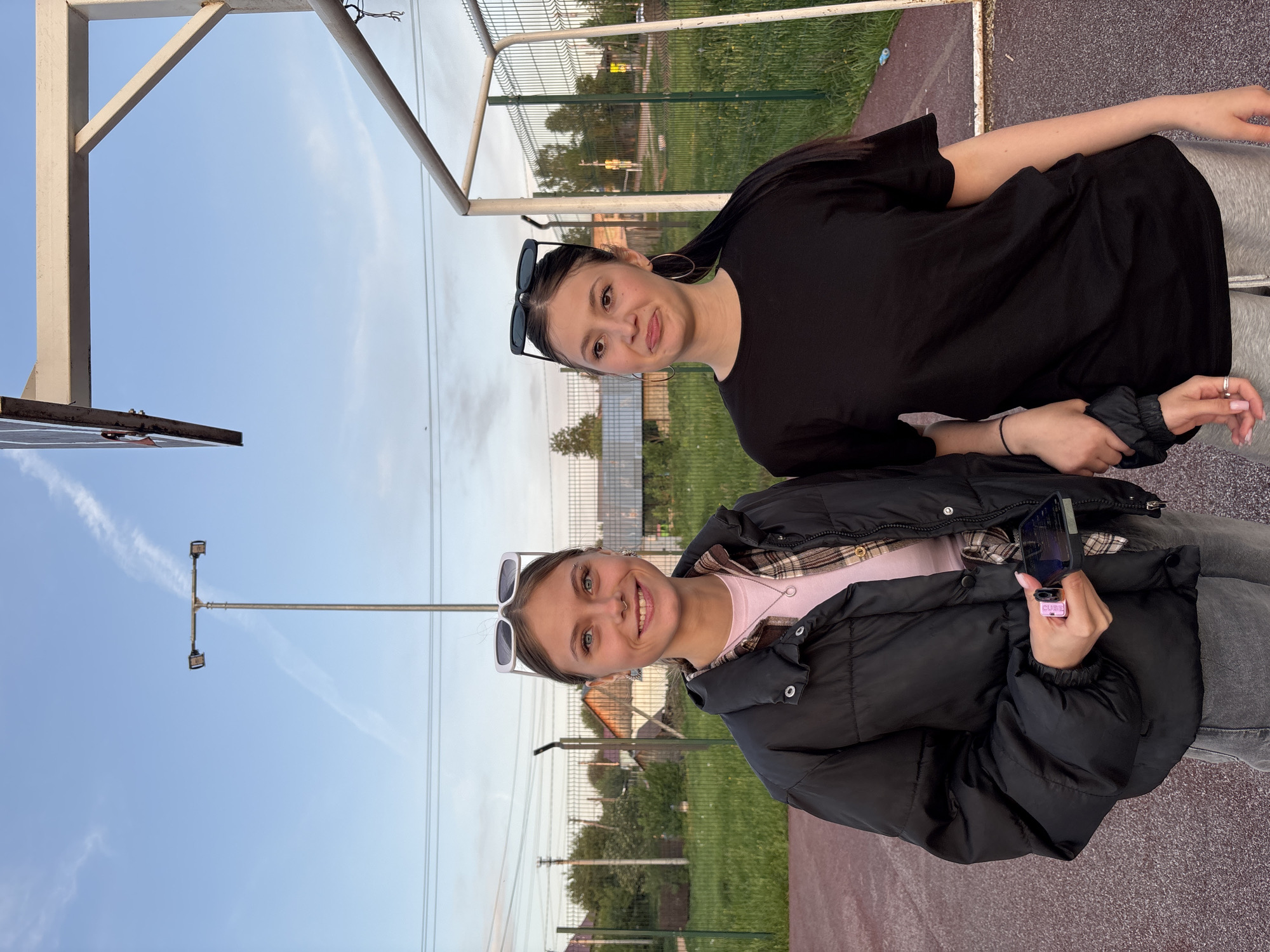 Two young women are posing outside in Фоки, Russia. The woman on the bottom is wearing a black puffer jacket over a pink shirt and gray jeans, with white sunglasses on her head. She is holding a phone with a pink case. The woman on the top is wearing a black dress and has black sunglasses on her head. They are standing near a metal fence and a basketball hoop. The sky is blue with some clouds, and there is a streetlight pole in the background.