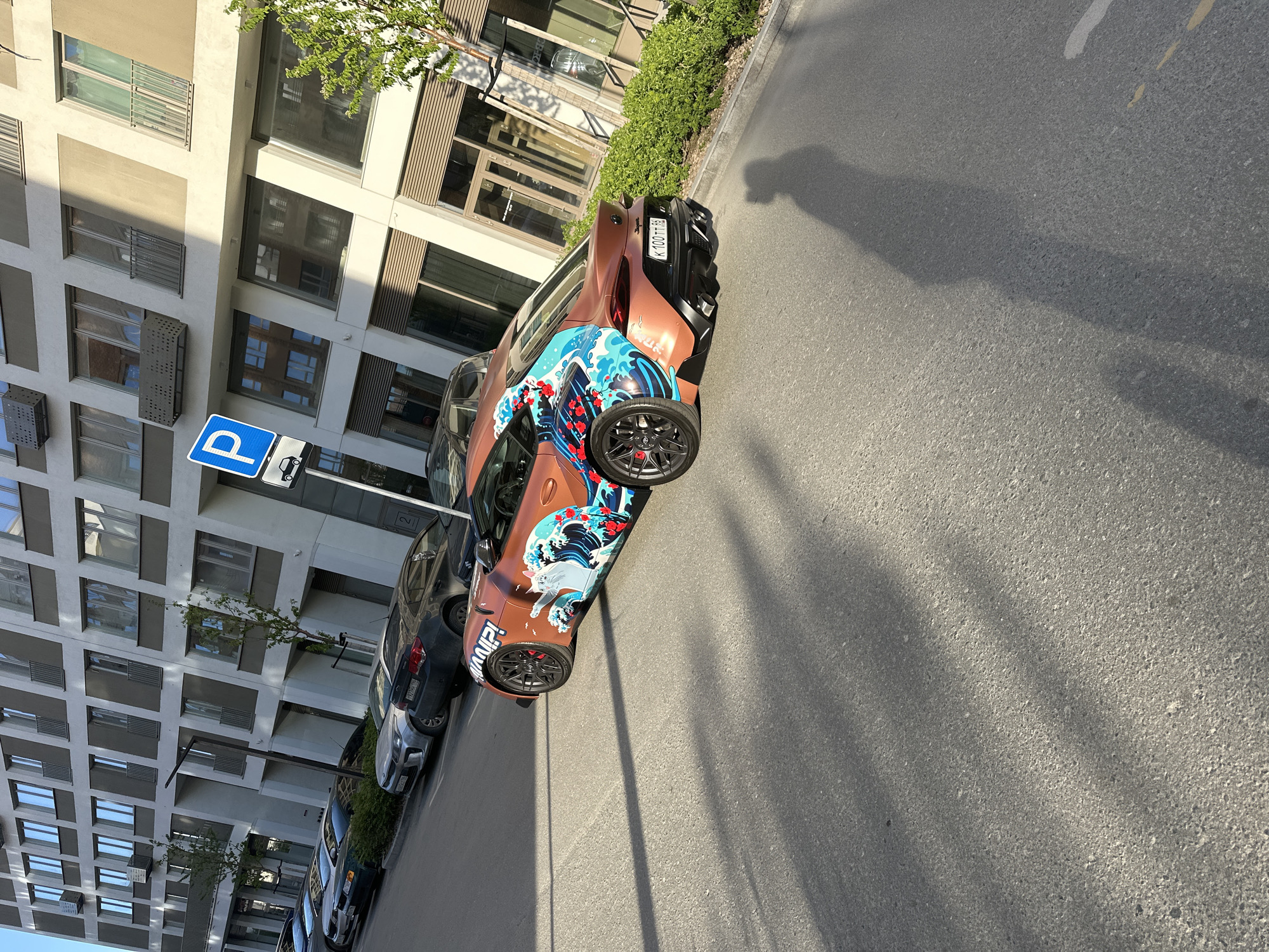 The image depicts a street scene in Yekaterinburg, Russia, featuring a uniquely designed car parked along the side of the road. The car is predominantly copper-colored, with a large, eye-catching graphic of a stylized wave and floral elements adorning its side. The car’s license plate is visible, reading "K 100 TT 96." The vehicle appears to be parked in a designated parking zone, as evidenced by the blue parking sign adjacent to the parked cars. The backdrop consists of a multi-story building with numerous windows and a tree growing on the side. Additional parked vehicles are visible in the background, and a shadow stretches across the street. The overall lighting suggests it is daytime.