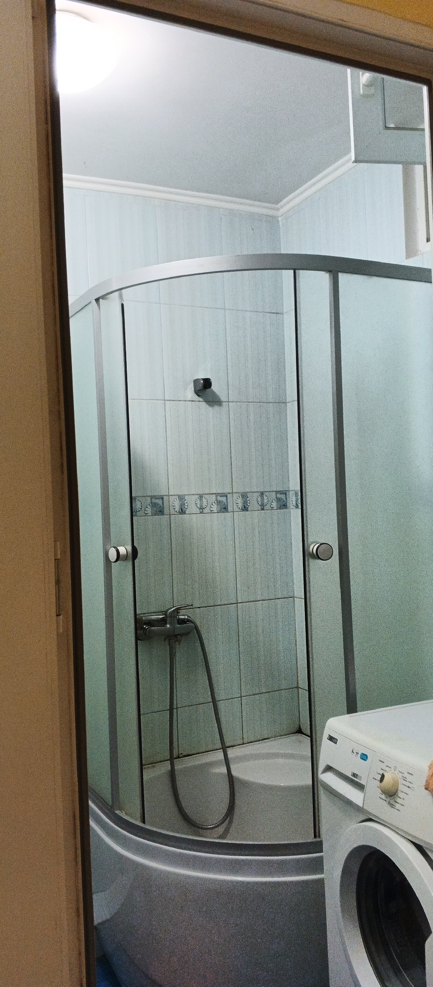 The image shows a corner shower stall with frosted glass doors and a white bathtub. The walls inside the shower are tiled with light blue tiles and a decorative border. A showerhead and hose are visible. To the right of the shower, a white washing machine is partially visible. The scene appears to be in a bathroom. The image is an interior shot.