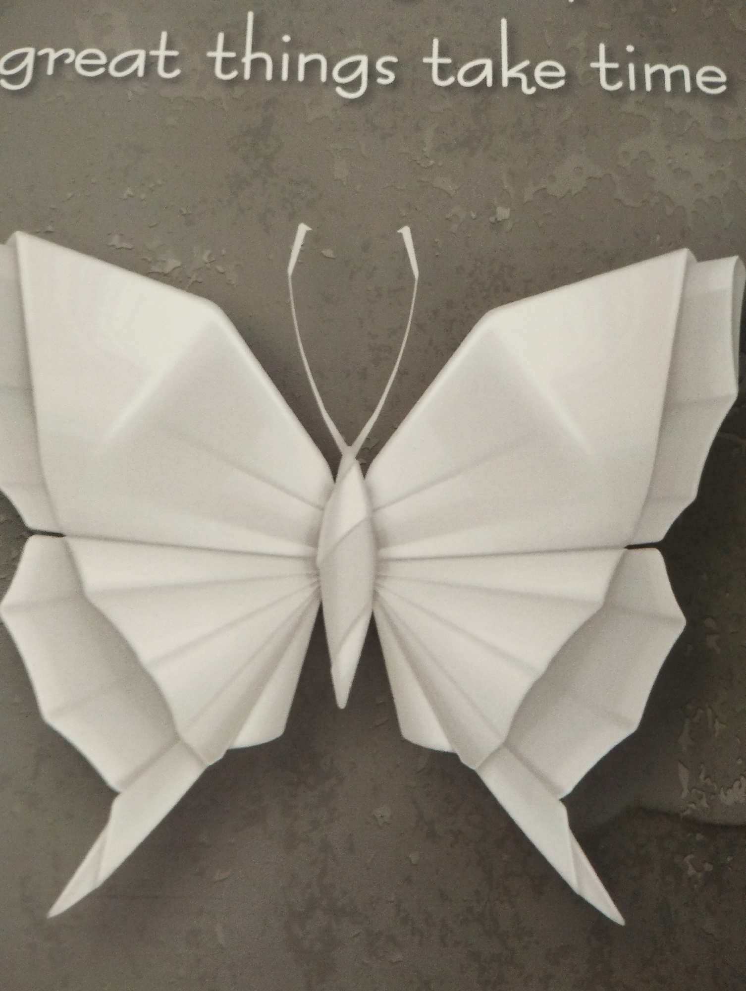 This media file features a close-up, overhead view of a white origami butterfly. The butterfly is centrally placed against a textured, gray background that appears to be a wall, possibly with some peeling paint or a concrete finish. The butterfly's wings are intricately folded, creating sharp angles and a sense of depth. Two thin, white antennae extend upwards from the butterfly's body, curving slightly.
Above the butterfly, white text is visible against the gray background. The text is in a cursive font and reads: "great things take time".
The lighting suggests it is indoors or during daytime, as there are no strong shadows indicating a specific time of day. The overall mood conveyed is calm and inspirational, due to the subject matter and the motivational text. The setting appears to be a wall in a domestic or public space in Pardes Hana-Karkur, Israel, likely a decorative element. No people or specific events are depicted.