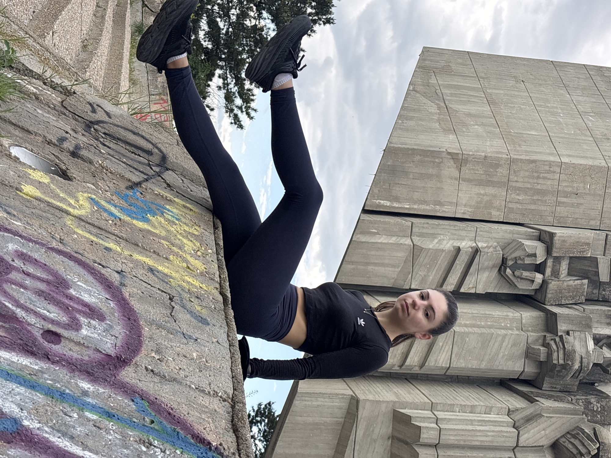 A woman in black athletic wear poses on a large, concrete structure in Varna, Bulgaria. She wears a long-sleeved crop top with an Adidas logo, leggings, and black sneakers. Her legs extend upward towards the sky, and she looks directly at the camera. The structure has graffiti on one side and is made of large concrete blocks. The sky is cloudy, and trees are visible in the background.