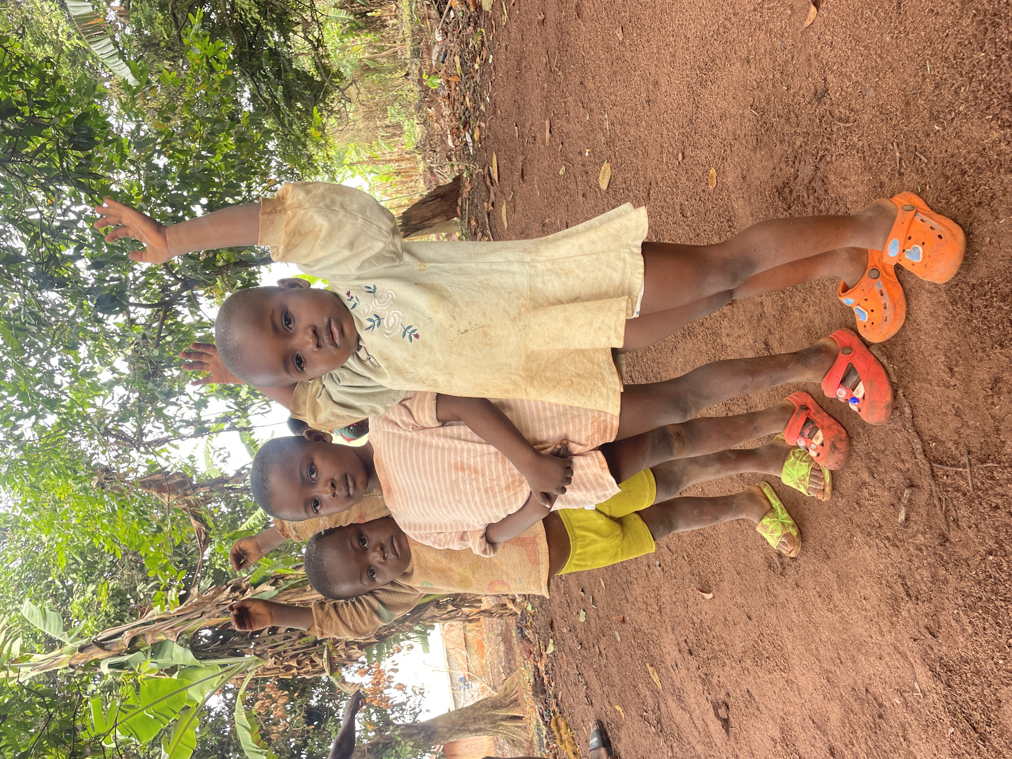 Three children are standing in a line, one behind the other, on reddish-brown dirt in Ogbe, Nigeria. The background consists of lush green foliage and trees. The first child is wearing a cream-colored dress and orange sandals, the second child is wearing a striped peach shirt and red sandals, and the third child is wearing a brown shirt and yellow shorts with yellow sandals. All three children are facing forward with their arms raised.