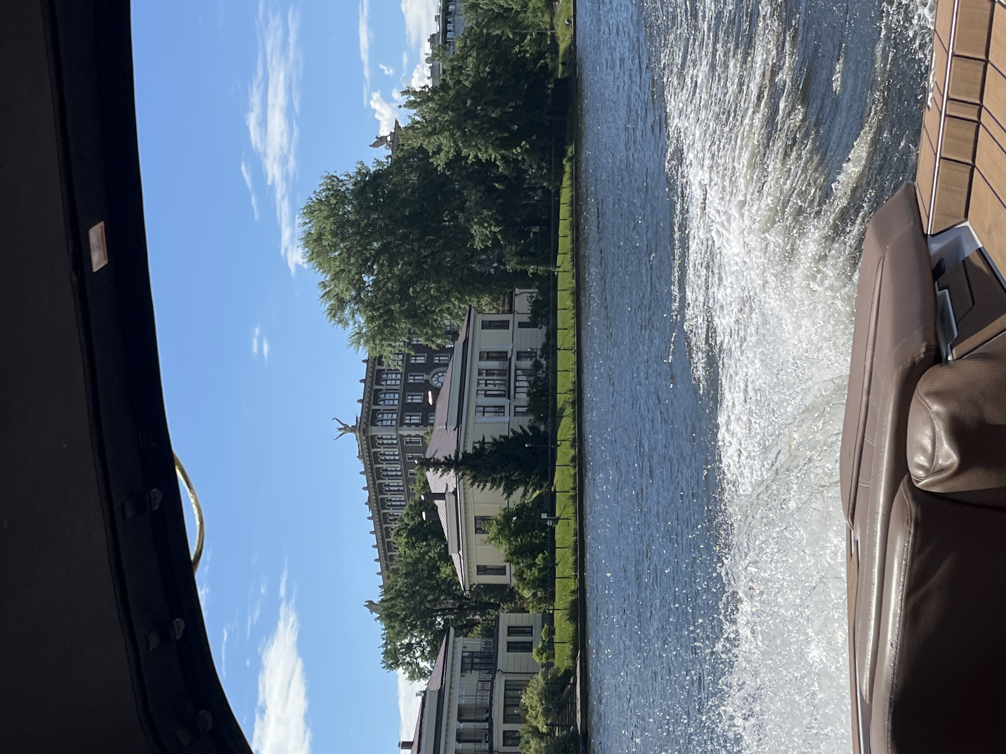 A view from a boat on a sunny day, looking towards the Petrograd Side of Saint Petersburg, Russia. The water is dark blue with white wake. On the shore, there are several modern buildings with classical architectural elements, including balconies and columns. Green trees and manicured lawns are visible between the buildings. In the background, a larger, darker building with multiple windows and decorative cornices stands prominently, topped with sculptures of winged figures. The sky is bright blue with scattered white clouds. The foreground shows the brown leather interior of the boat.