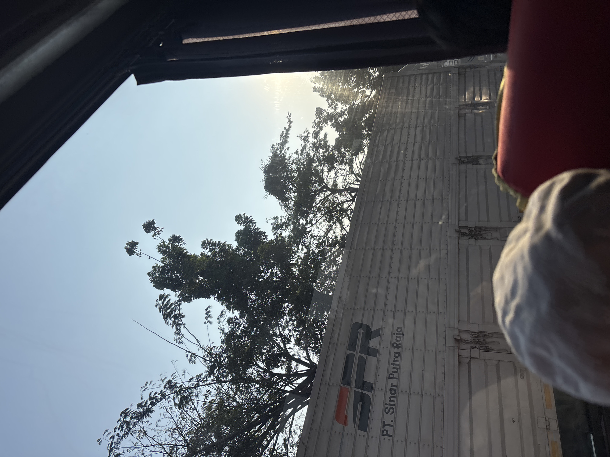The image shows a section of a large, ribbed metal truck trailer, likely in motion, viewed through a window. The trailer is light grey and displays a black and orange logo with the letters "SPR" and the text "PT. Sinar Putra Raja". Above the trailer, silhouetted trees with dense foliage are visible against a clear blue sky. Sunlight filters through the trees and reflects off the truck's surface, creating glare. In the foreground, partially in focus, is the back of a person's head wearing a light-colored cap, and a section of a red seat, suggesting the photo was taken from inside a vehicle. The overall impression is a snapshot of travel, likely on a road in Hegarmukti, Indonesia, given the context.