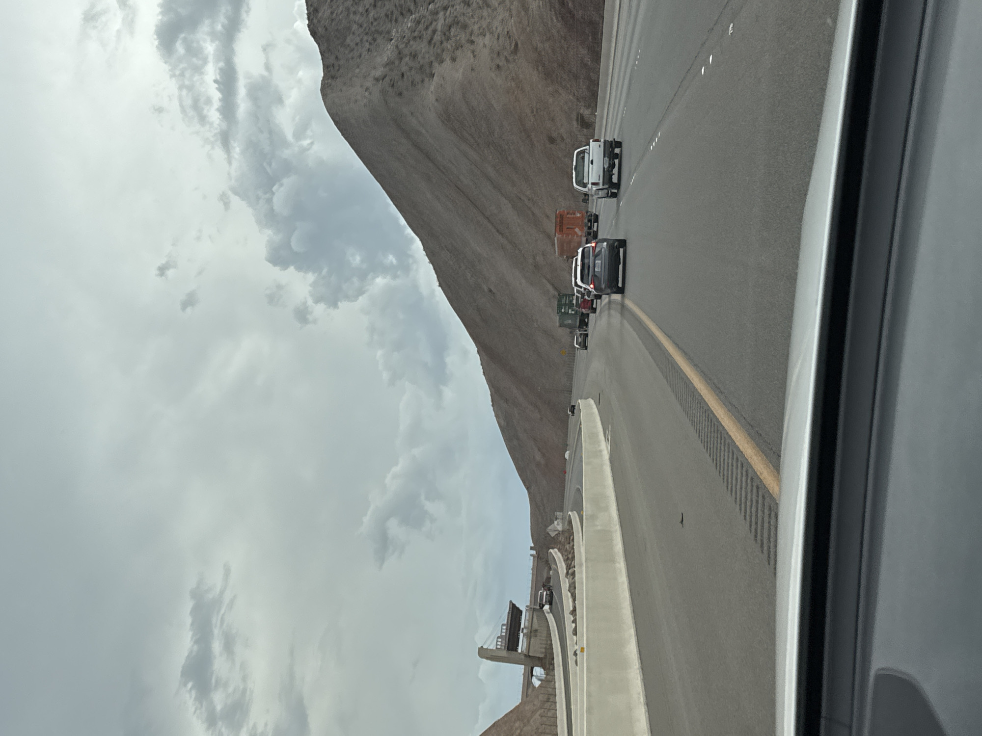 The image depicts a view from a vehicle traveling on a highway in Boulder City, Nevada, USA. The highway curves to the right, bordered by a concrete barrier on the left and a mountainous desert landscape on the right. The sky is overcast with dramatic clouds. Several vehicles are visible on the highway, including cars and trucks. In the distance on the left, a bridge structure is under construction, with its support pillars visible. The overall scene suggests a journey through a rugged, arid environment.