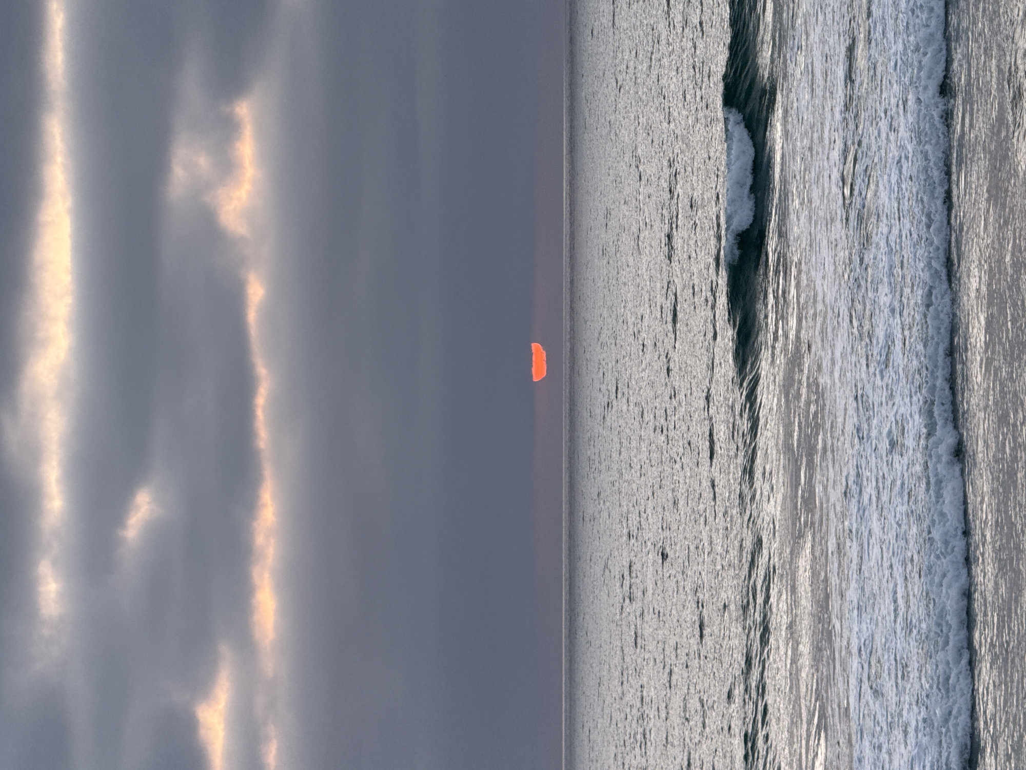 The image captures an ocean sunset scene. The right half of the frame is dominated by the ocean, with visible waves and white foam. The left half consists of a cloudy sky, with streaks of light breaking through. In the center, a small, vibrant orange sun sits on the horizon line. The scene appears serene and natural. The location is plausibly the city of San Diego, United States of America, since the city faces the Pacific Ocean and is known for its sunsets. There are no identifiable people, subjects, objects, actions, or interactions besides the natural elements of the sky, ocean, and sun.
