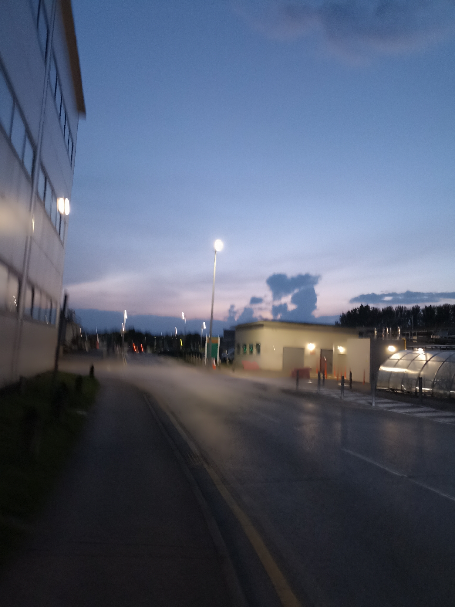 The image depicts an outdoor scene at dusk, likely in an industrial or commercial area. A modern building with a white facade and multiple windows is visible on the left. A paved road, possibly a street or access road, runs through the center of the frame, with yellow lines marking the lanes. Mist or fog is present over the road, creating a hazy effect.
Several streetlights are illuminated, casting a warm glow. In the background, a low-lying building with lights on is visible, along with what appears to be a covered walkway or structure with reflective panels. Trees line the distant horizon. The sky is a gradient of blue and purple, characteristic of twilight.
Based on the context provided by the prompt mentioning the city of Galway, Ireland, this scene could be located in an industrial park or a business campus within or on the outskirts of the city. However, without specific landmarks or signage, it is impossible to pinpoint an exact location within Galway. The overall atmosphere is one of a quiet, functional environment transitioning from day to night.