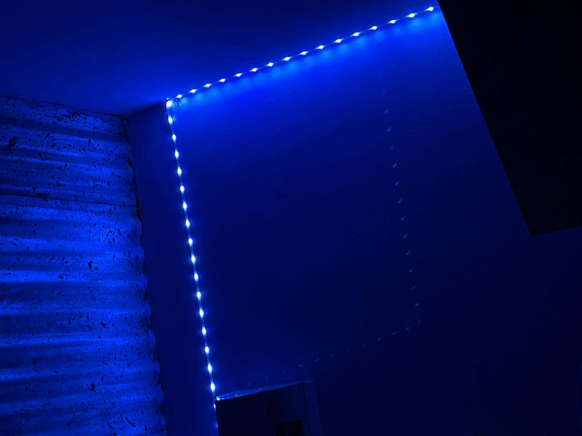 The image depicts the corner of a room illuminated by a blue LED light strip. The strip runs along the ceiling and down the wall, casting a blue glow on the surfaces. The wall to the left has a textured, layered appearance. A darker, undefined shape appears in the upper right corner, possibly part of the ceiling or an object in the room. There's a faint impression or shadow that might be a rectangular outline on the right-side wall. The lighting creates a stark contrast between illuminated areas and those in shadow. There is no evidence to support the claim that the location is in Juan Griego, Venezuela.