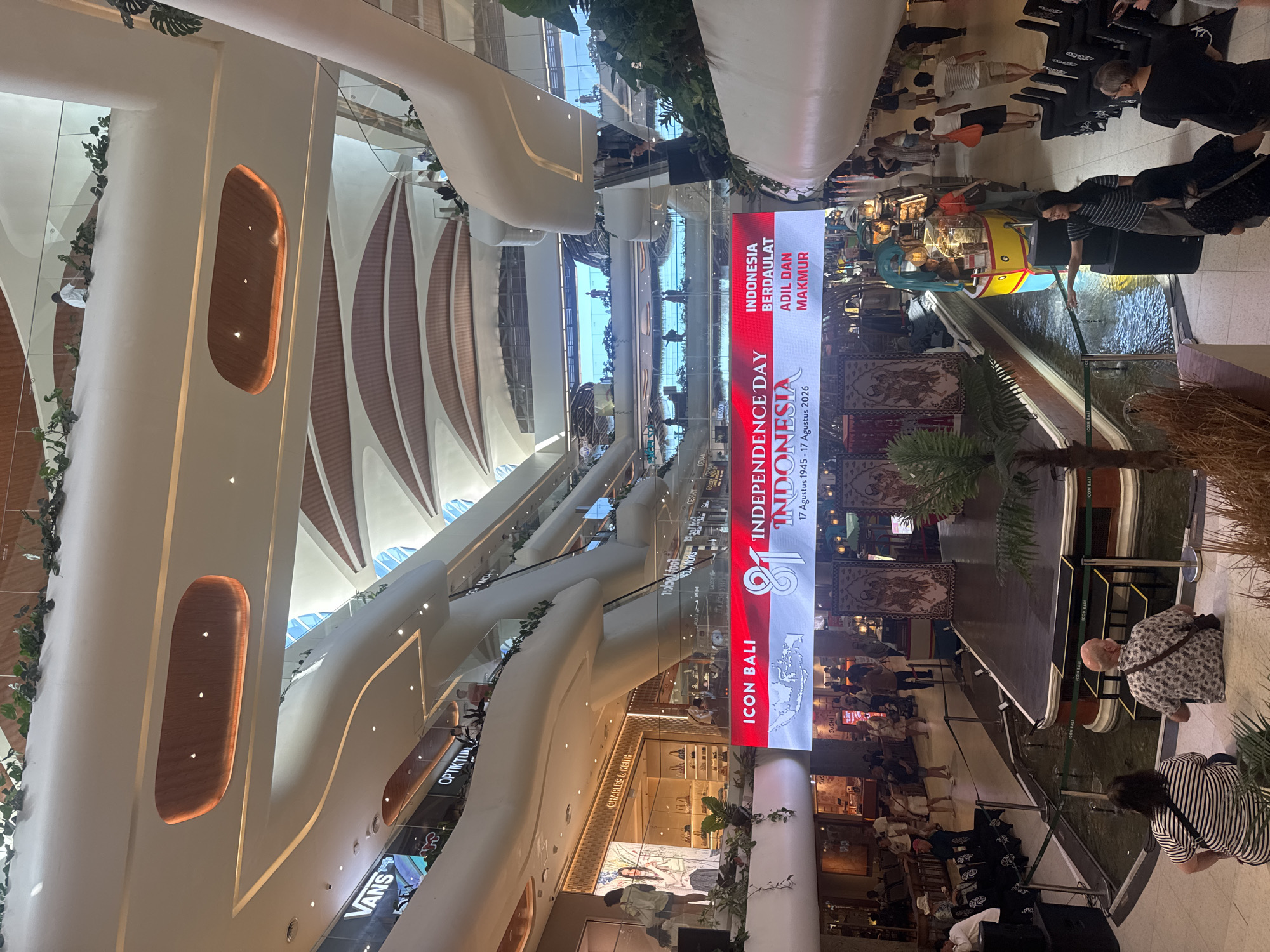 The image depicts the interior of a modern, multi-level shopping mall, likely in Denpasar, Indonesia, given the visible "ICON BALI" branding on a large LED screen. The screen prominently displays "INDEPENDENCE DAY INDONESIA" along with the Indonesian flag and dates "17 Augustus 1945 - 17 Agustus 2026," indicating a national celebration. Below the screen, a stage area is set up with traditional Balinese tapestries, suggesting a cultural performance or event.
The mall's architecture features organic, curved lines, with white and light wood finishes and abundant greenery integrated into the design. Escalators and walkways are visible on multiple levels, populated by a moderate number of shoppers and visitors. Several storefronts, including "VANS" and "OPTIC TUNK," are visible in the background. In the foreground, two individuals are seated on the floor, facing away from the camera, while others stand and walk around the central area, which includes a water feature and a small boat-shaped stall. The lighting suggests it is daytime, with natural light filtering in from upper levels.