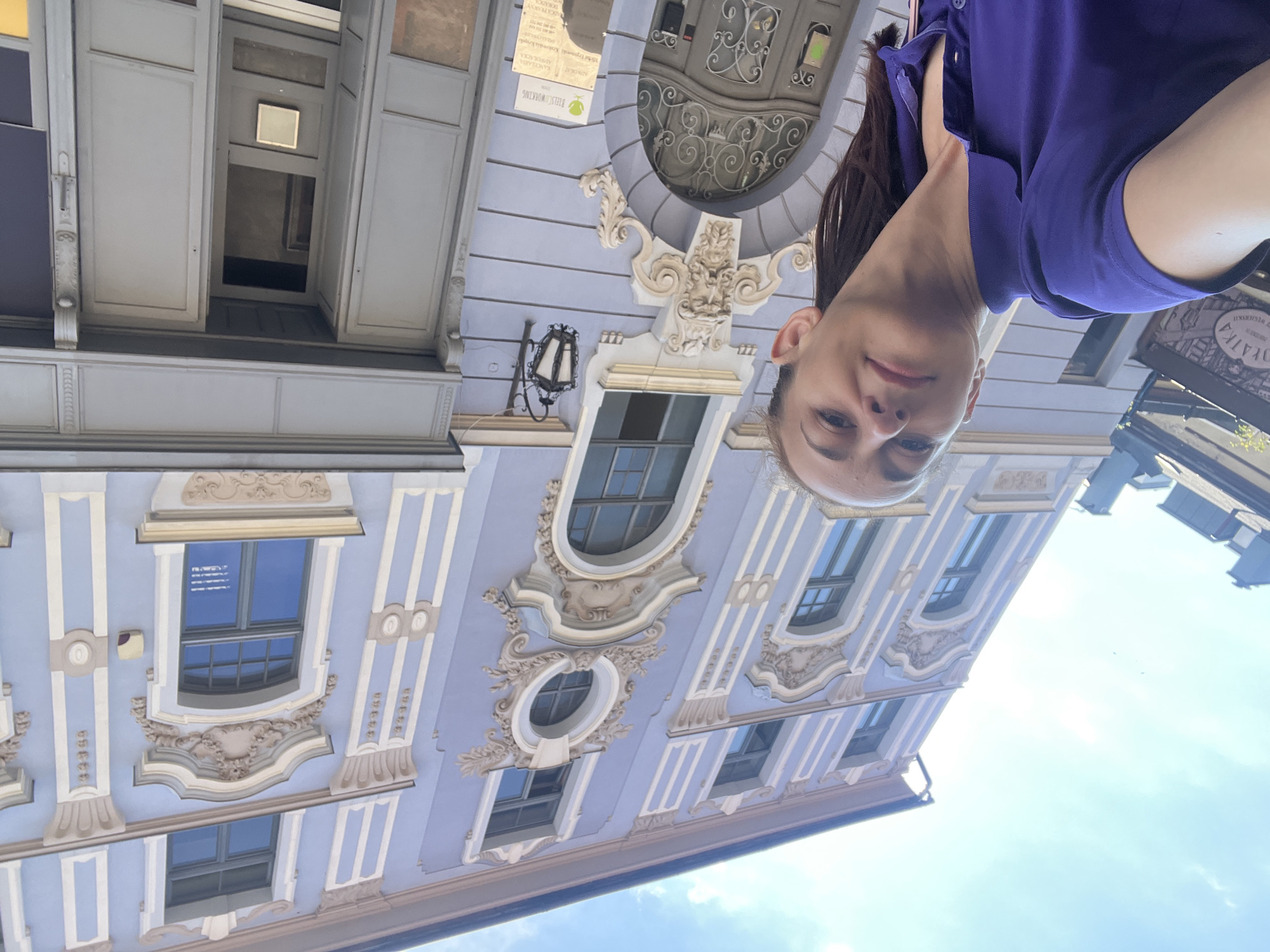 A woman is taking a selfie outdoors, leaning back against a light blue building with ornate white and beige architectural details. The building has several windows, some of which are arched. A black wrought-iron lantern hangs from the building. The woman is wearing a purple polo shirt. Part of a sign with text is visible on the building to the right of the woman. The sky above is blue with some clouds.