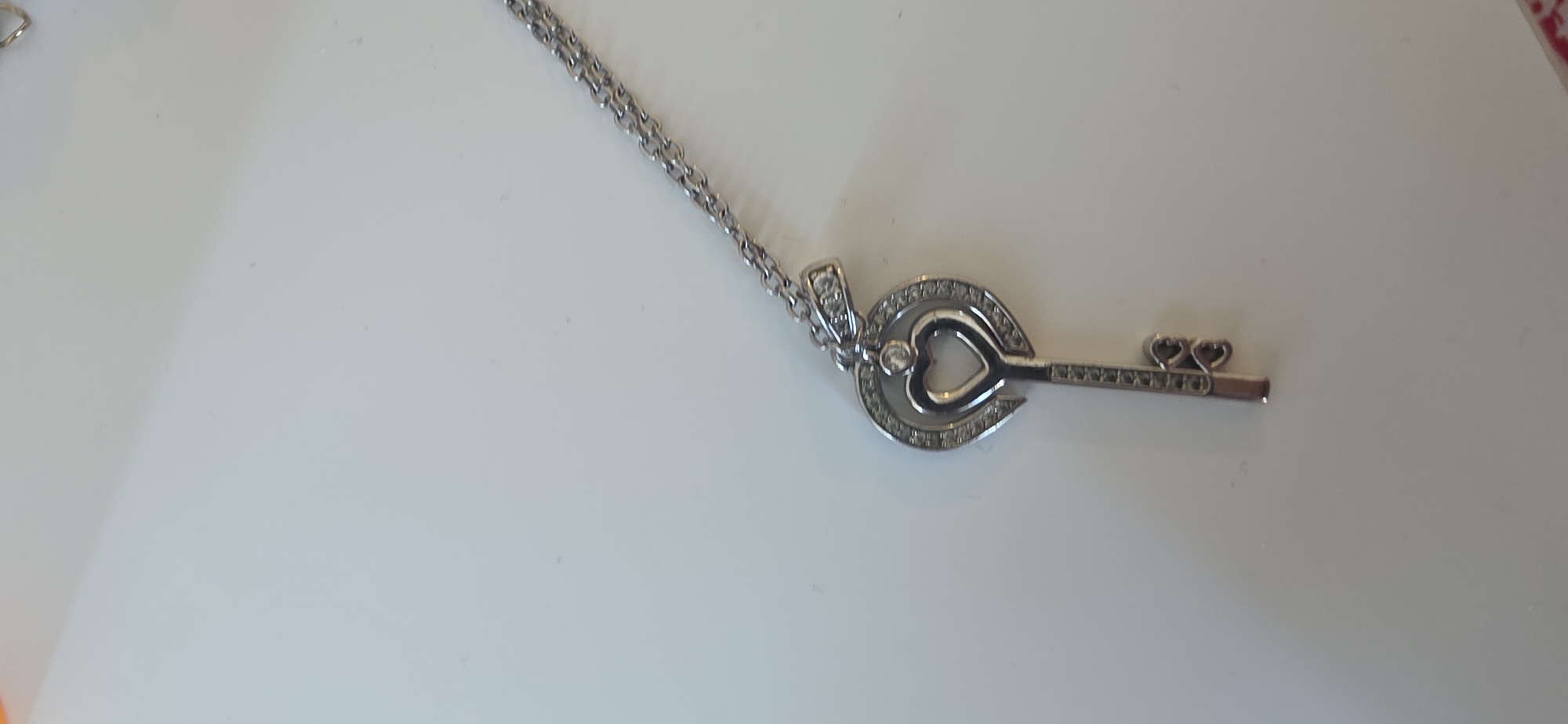 A close-up, high-angle shot shows a silver-colored necklace with a key-shaped pendant on a white, reflective surface. The pendant is intricately designed with a heart shape at the top, surrounded by small, clear stones. The key's shaft is also adorned with small stones, and the end of the key features two small heart shapes. The chain of the necklace is thin and metallic. The background is a plain white surface, likely a table or desk, with some faint shadows or reflections. There are no people or other objects visible in the frame. The image appears to be taken in a studio or indoor setting, with artificial lighting. The specific location within the city of Sumgayit, Azerbaijan is not discernible from the provided image.