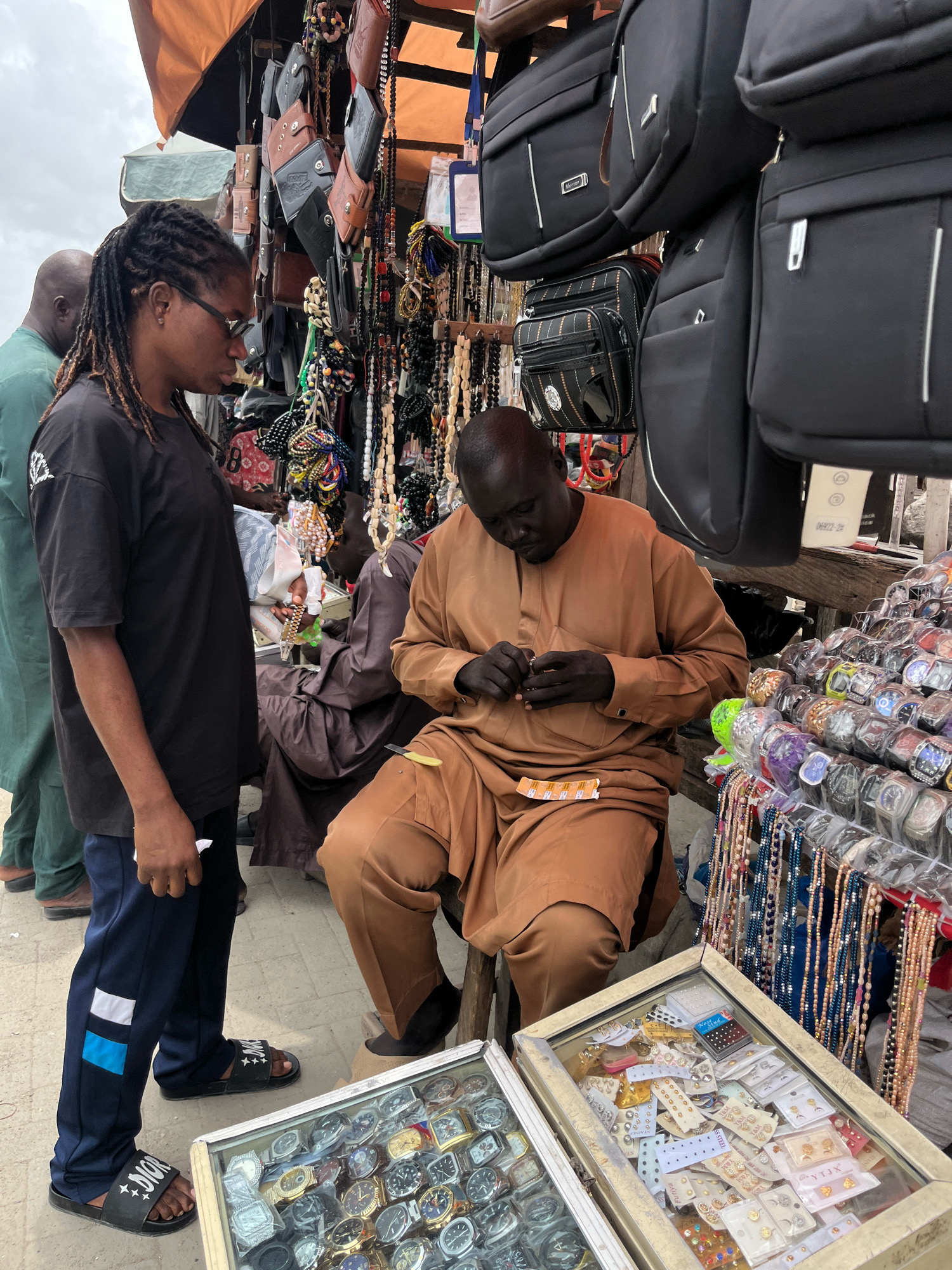 In Ajah, Nigeria, a street market scene unfolds. A woman with dreadlocks and glasses stands to the left, wearing a black t-shirt and dark pants. She is looking towards a man seated on a stool, who is wearing a brown tunic and trousers. The man is examining a small object in his hands. In front of him, a display case is filled with an assortment of wristwatches. Behind the man, a stall is laden with various bags, wallets, and numerous beaded necklaces and bracelets hanging from racks. Another person in a green garment is visible in the background to the left. The ground appears to be paved.