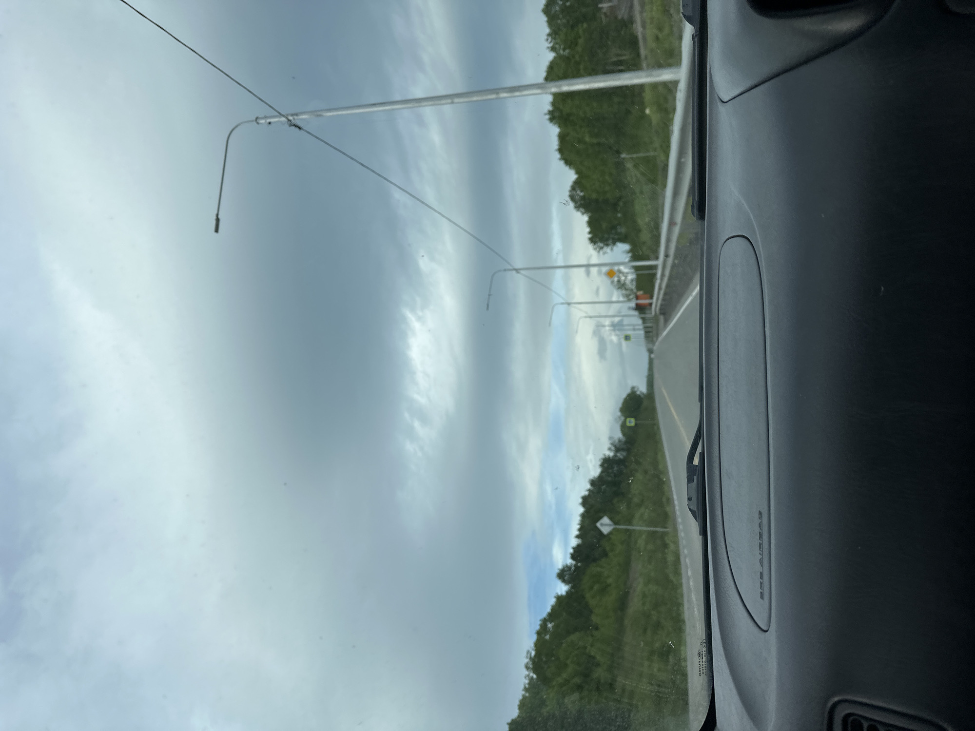 The image is a first-person perspective from inside a vehicle, looking out through the windshield at a rural road. The sky is overcast with heavy gray clouds, with a small patch of blue sky visible in the distance. On either side of the road are dense green trees and foliage. Several streetlights are visible along the road, spaced at intervals. A guardrail runs along the right side of the road. The road itself is paved and appears to be a two-lane highway. In the foreground, the dashboard of the vehicle is visible, with the text "SRS AIRBAG" embossed on it. The location is identified as being in the city of Svetly, Russia.