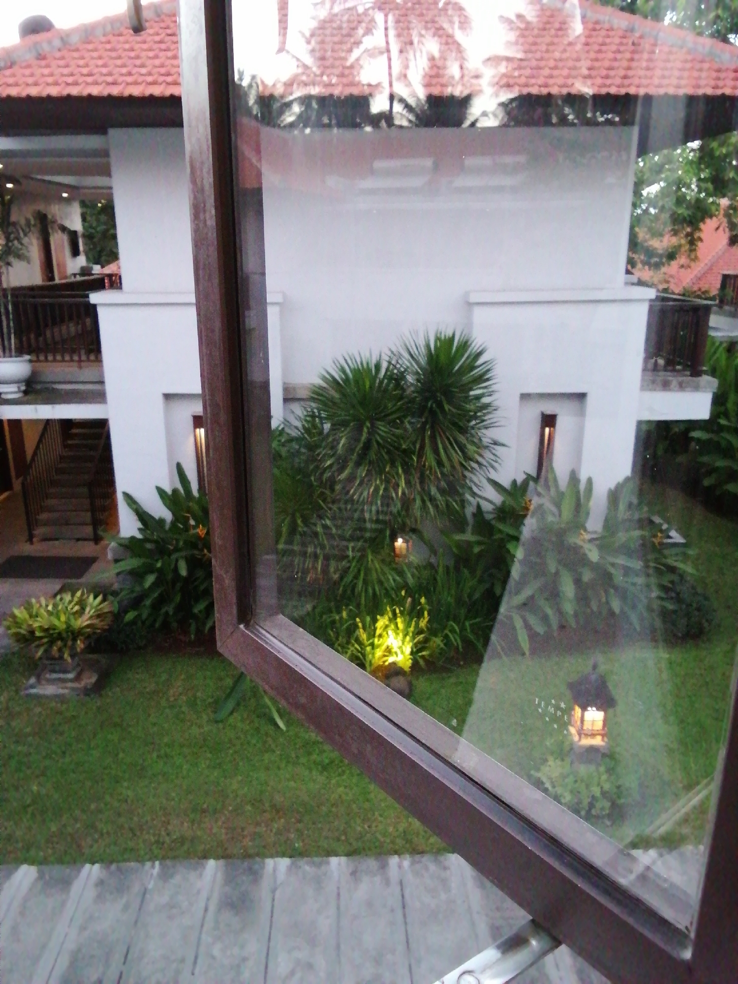 The image shows a view through a window onto a courtyard with lush green grass and tropical plants. The courtyard is flanked by a white building with a terracotta tiled roof and dark wooden railings. A set of stairs with dark railings leads up to the building. In the distance, a small, illuminated lantern sits amongst the foliage. The scene suggests a tranquil, possibly resort-like, setting. The window frame, made of dark brown wood, is partially open, indicating the viewpoint is from inside a building looking out. The overall impression is one of peaceful outdoor living.