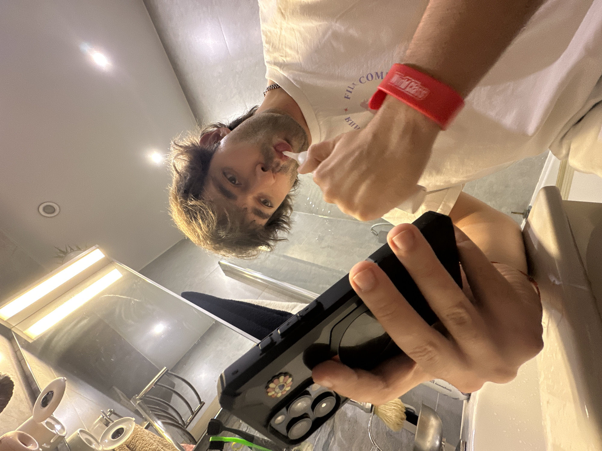 A young adult male is seen in a bathroom, possibly in Moscow, Russia. He is holding a smartphone in his left hand and brushing his teeth with his right hand. He is wearing a white t-shirt with red text on it and a red wristband. The bathroom appears to be clean and modern, with a sink, mirror, and shower visible. The lighting is bright, and the overall impression is one of a casual, everyday activity.