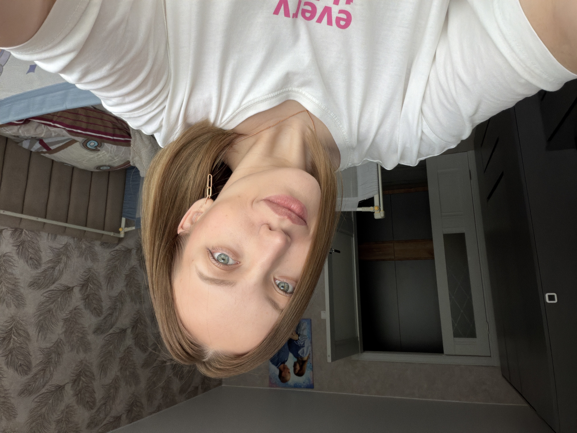 The image shows a selfie of a person with light skin, light eyes, and light brown hair. They are wearing a white t-shirt with pink lettering. A gold chain and earrings are visible. The background includes a bed with patterned bedding, a patterned wall, a grey cabinet, a closed white door, and a picture of two people hanging on the wall. Based on the prompt, it is presumed that the location is in Reshetnikovo, Russia.