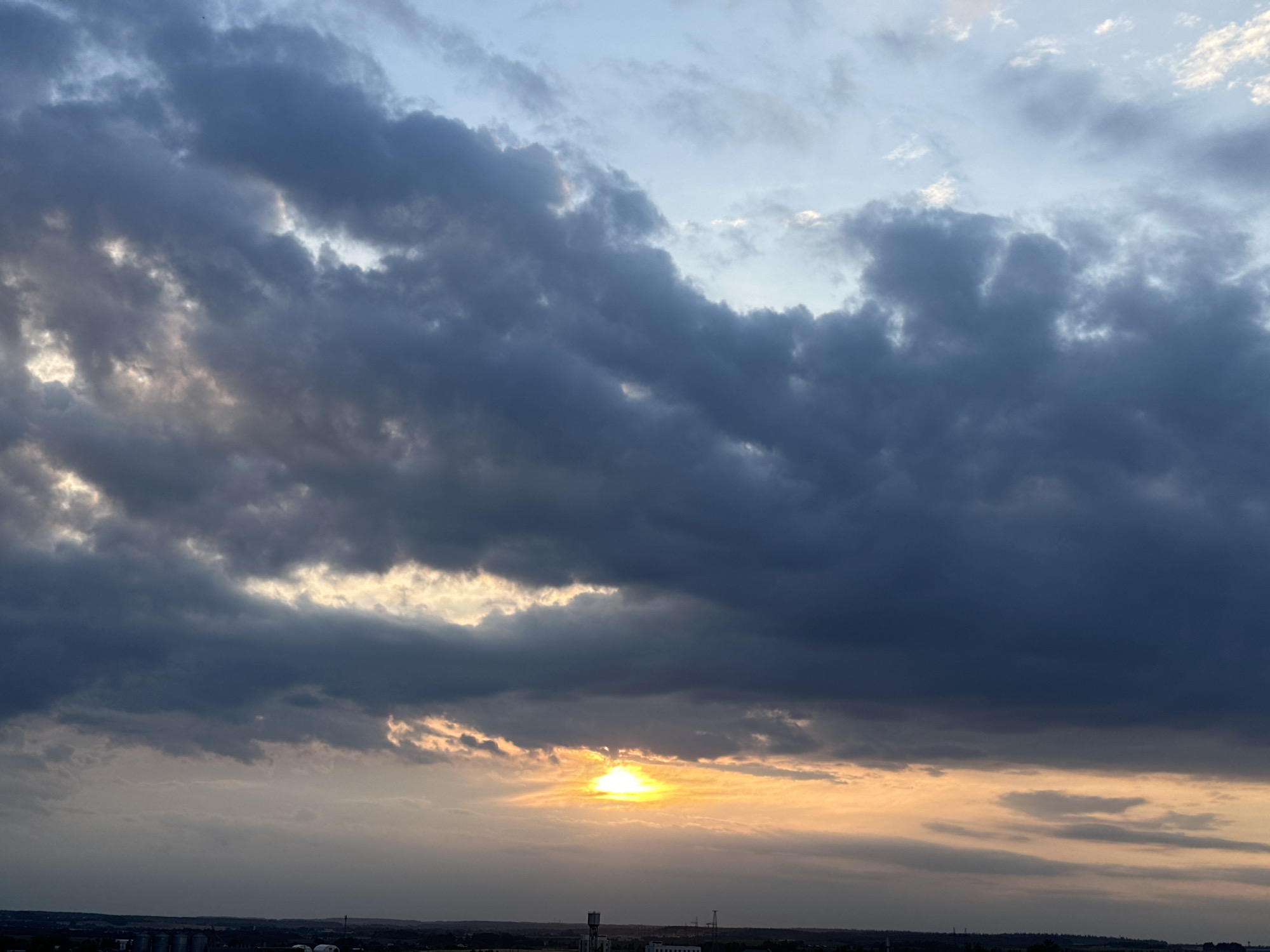 The image depicts a dramatic sky over the city of Shchomyslitski Selski Sovet, Belarus. Dark, heavy clouds dominate the upper portion of the frame, creating a sense of impending weather. Towards the horizon, the clouds break to reveal a vibrant sunset, casting an orange and yellow glow. The sun itself is a bright, concentrated orb peeking through a gap in the clouds. Below the sky, a silhouetted cityscape is visible, featuring various industrial structures and buildings, indicative of an urban or semi-urban environment. The lighting suggests it is either sunrise or sunset, with the low angle of the sun creating long shadows and a dramatic contrast between light and dark. There are no people or specific actions depicted. The scene is a natural landscape focused on the sky and the urban horizon.