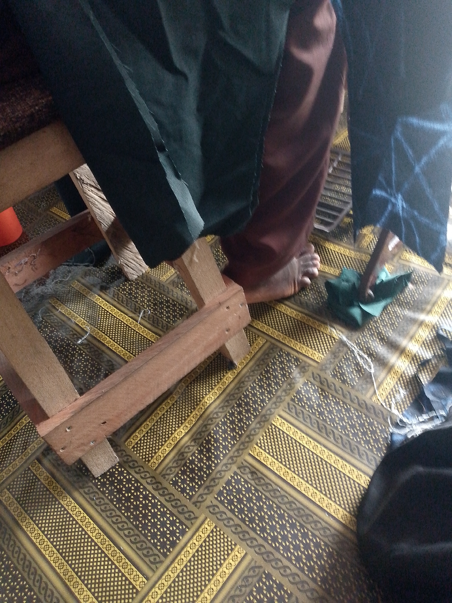 A low-angle, indoor shot captures a scene with a person's legs and feet on a patterned mat. The person is wearing dark green and brown garments. To the left, a wooden frame structure is visible, possibly part of a chair or table. The mat features a geometric pattern in yellow and dark brown. In the foreground, a small folded piece of green fabric lies on the mat, and a thin white string is also present. To the far right, a dark rounded object, likely a hat or a bag, is partially visible. The environment appears to be in Jauro Yinu, Nigeria, suggesting a domestic or workshop setting.