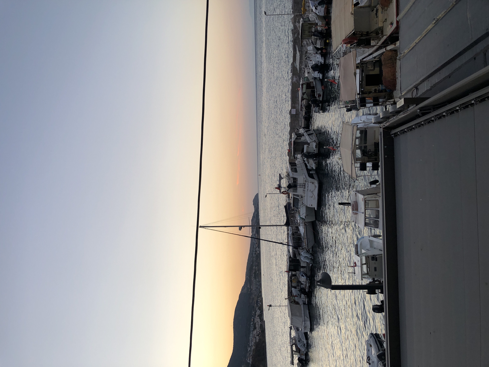 The image shows a harbor filled with boats at sunset in Karaburun, Turkey. The sky is a gradient of pale blue to soft orange, indicating the sun is low on the horizon. The sea is calm and reflects the colors of the sky. In the foreground, a dark, flat surface, likely a pier or rooftop, occupies the bottom of the frame. Behind this, a row of moored boats of various sizes and designs fills the water. Some are small, open boats with outboard motors, while others are larger cabin cruisers and a sailboat with a prominent mast. The boats are docked along a rocky breakwater, and a few appear to be fishing vessels with nets visible. The coastline of Karaburun is visible in the background, with hills and buildings stretching along the shore under the warm glow of the setting sun. A single black wire stretches across the upper portion of the image, adding a linear element to the scene. There are no people visible in the image.
