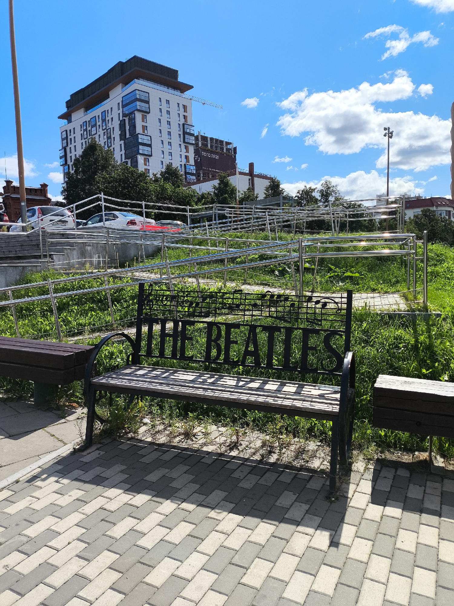 This image shows a park bench with the inscription "THE BEATLES" on its backrest. The bench is located in Yekaterinburg, Russia, in front of a modern residential building. The area is paved with gray and beige tiles, and there is green grass and trees around. In the background, there are cars, a ramp, and a construction crane. The sky is blue with white clouds.