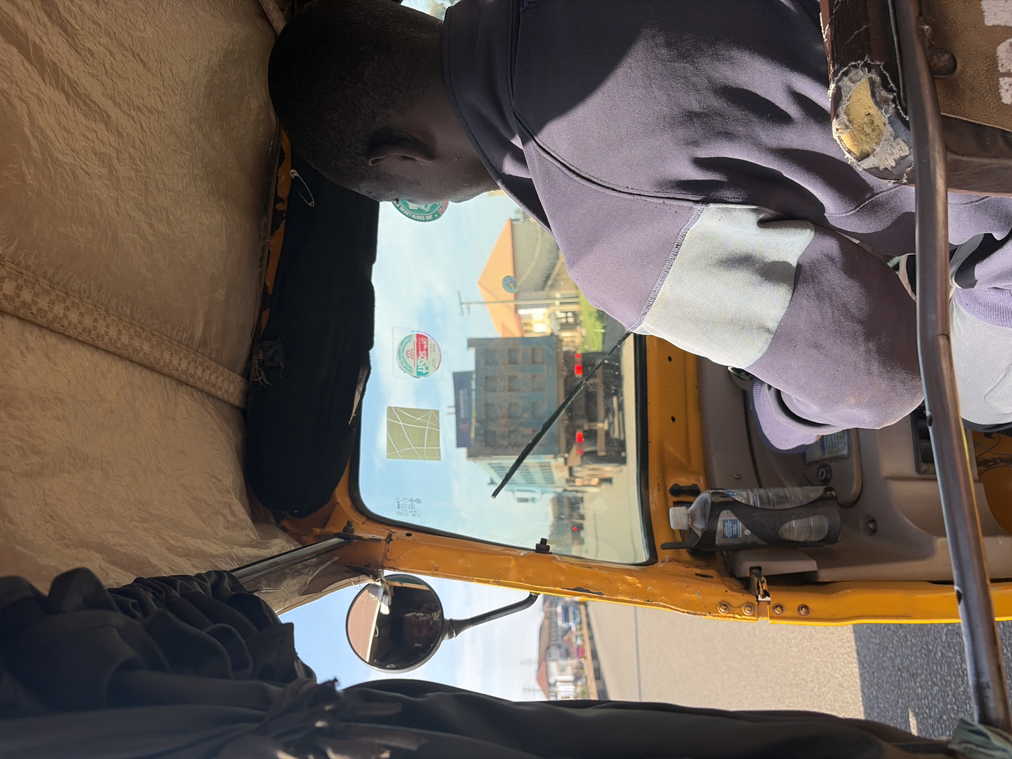 The image shows a view from inside a yellow auto-rickshaw driving on a road, likely in the city of Gura Top, Nigeria. The driver, a Black man, is facing forward, wearing a dark purple jacket with lighter purple panels. He is looking out through the windshield. In the reflection of the side mirror, a child is visible, appearing to be smiling. Through the windshield, a large blue truck is visible ahead, along with other vehicles and buildings in the distance. The interior of the auto-rickshaw is visible, including the dashboard, a water bottle holder with a water bottle, and worn upholstery on the passenger seat. The sky is blue with scattered clouds.