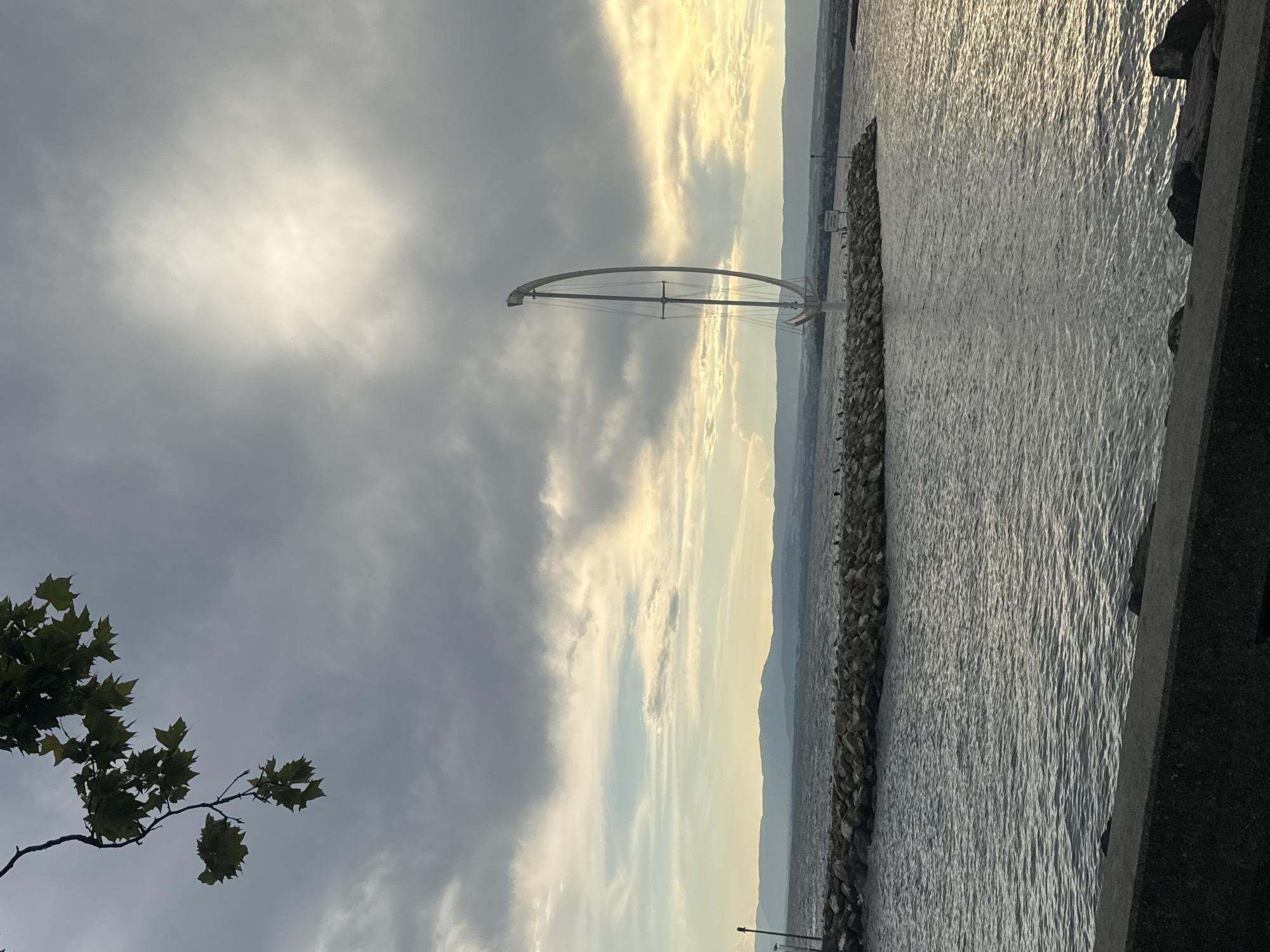 The image shows a view of Lake Geneva from the Ouchy district of Lausanne, Switzerland. In the foreground, a concrete embankment separates the viewer from the water. The lake is choppy, with sunlight reflecting off the surface. A jetty made of large rocks extends into the water, with several birds perched on it. At the end of the jetty stands a tall, abstract sculpture resembling a sailboat mast with a curved frame. In the background, a hazy mountain range is visible across the lake under a dramatic sky filled with dark clouds and streaks of sunlight. Overhanging branches with green leaves are visible in the upper left corner of the frame.