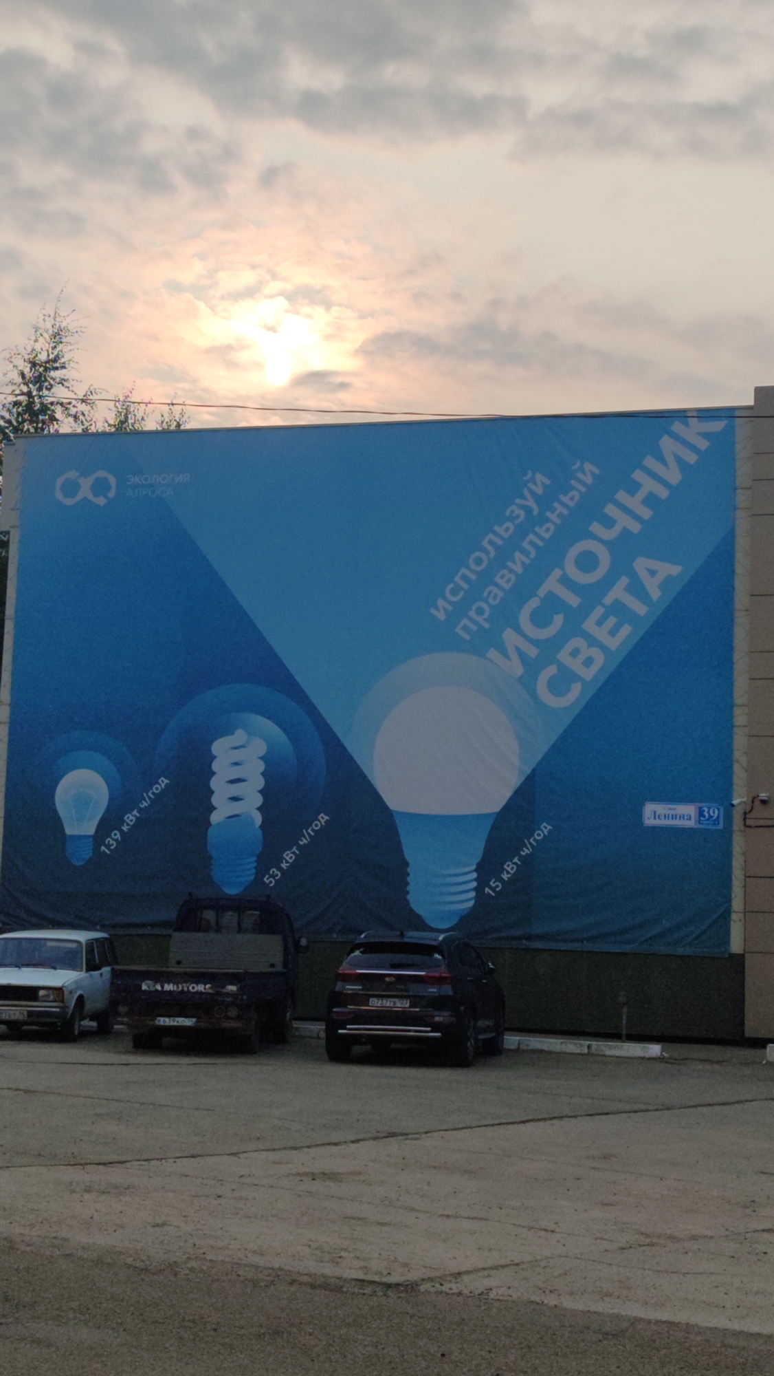 The image shows a large billboard in Mirny, Russia, promoting energy-efficient lighting. The billboard displays three light bulbs of varying designs, each with accompanying text indicating their energy consumption in kWh per year. The text on the billboard is in Russian, featuring phrases like "ЭКОЛОГИЯ АЛРОСА" (Ecology ALROSA) and "ИСПОЛЬЗУЙ ПРАВИЛЬНЫЙ ИСТОЧНИК СВЕТА" (Use the right light source). There is also a street sign that reads "Ленина 39" (Lenin 39), indicating the location.
In the foreground, several vehicles are parked: a white sedan on the left, a dark-colored pickup truck in the center with "KIA MOTORS" visible on its side, and a dark SUV on the right. The vehicles appear to be parked on an asphalt surface. The sky above is partly cloudy with the sun visible through the clouds, suggesting it might be late afternoon or early morning. The billboard is mounted on a building facade, and a security camera is visible to the right of the street sign. There are no people visible in the image.