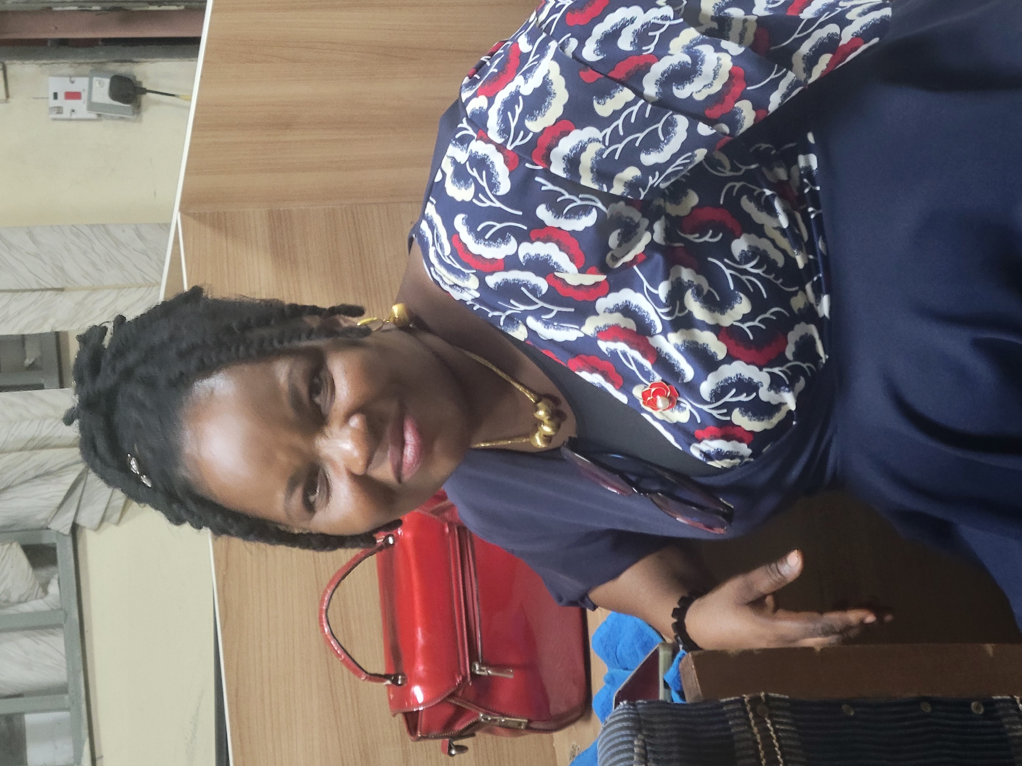 A woman stands indoors in Abuja, Nigeria. She has dark hair in dreadlocks and wears a navy blue dress, accessorized with a gold necklace, a bracelet, and a navy blue jacket with a white, red, and navy blue pattern. A red handbag sits nearby. An electrical outlet is visible on the wall behind her.