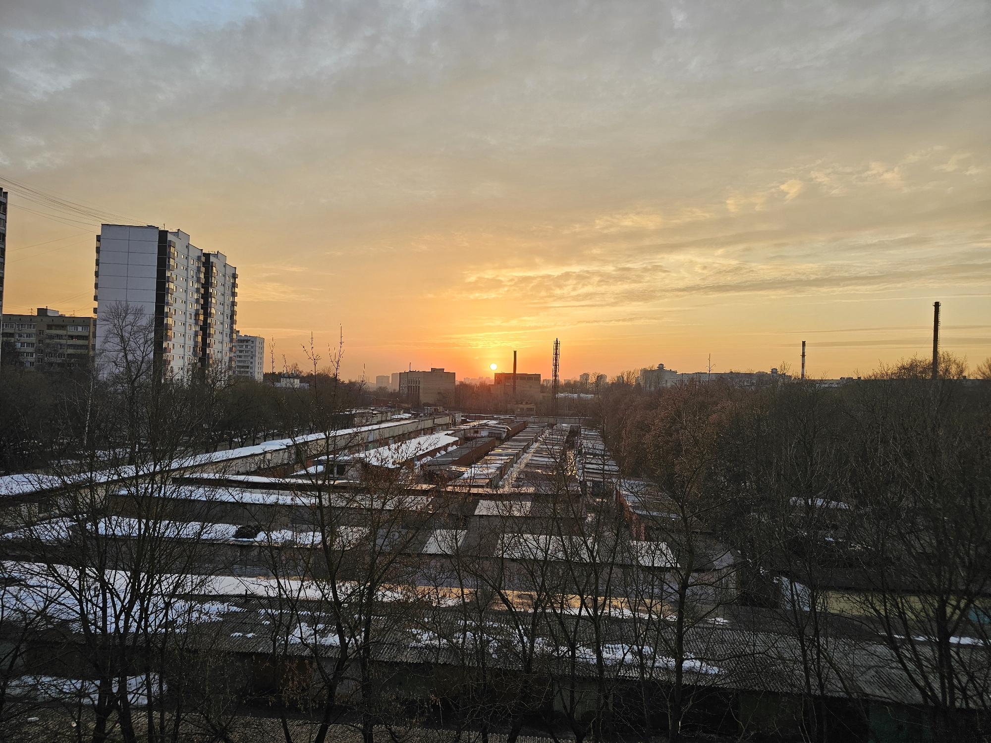 The image depicts an elevated view of an urban landscape in Khimki, Russia, at either sunrise or sunset during late winter or early spring. The sky transitions from a warm golden-orange hue near the horizon, where the sun is visible as a distinct orange orb slightly left of center, to muted grey and light blue clouds in the upper portion. In the immediate foreground, a dense network of bare deciduous tree branches partially obscures the scene below. The midground features an extensive complex of single-story garage structures, uniformly constructed with sloped roofs. Many of these roofs retain patches of residual snow and exhibit reflective wet surfaces, indicating thawing. To the left, several multi-story residential buildings are visible, including a prominent white and dark-paneled apartment block with overhead utility lines running past it. Further in the distance, beneath the low sun, an industrial area is present, marked by at least two prominent, dark, slender smokestacks or towers and other low-rise industrial buildings. A third smokestack is visible on the far right horizon. No individuals or specific activities are discernible in the static scene.