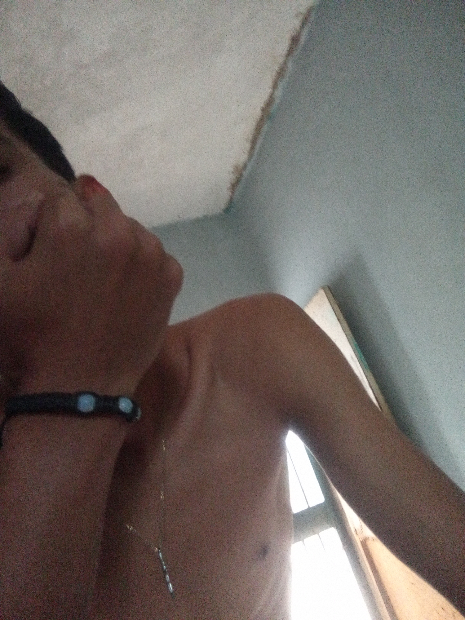 A dimly lit, low-angle indoor shot depicts a fair-skinned individual from the chest up. The person is shirtless, with a thin silver necklace and a black beaded bracelet visible. Their face is turned upwards, partially obscured. A window with bright light coming through is visible in the background, as are walls with uneven textures and pale paint. The image does not give information that can be used to confirm the location in the city of Juan Griego, Venezuela.