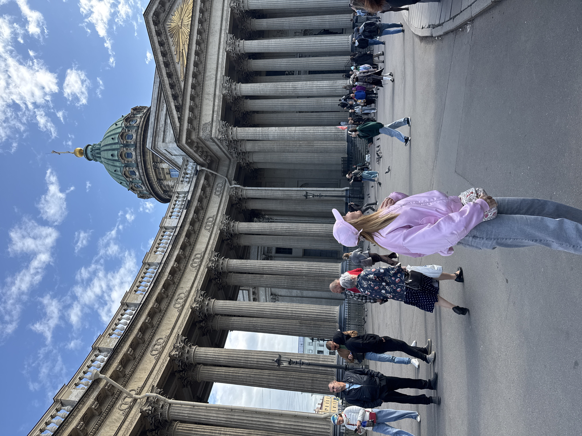 The image shows the Kazan Cathedral in Saint Petersburg, Russia. The cathedral, with its grand columns and dome, dominates the left side of the frame, reaching into a blue sky with scattered clouds. A number of people are walking in front of the cathedral, many of whom are dressed in casual attire. The scene appears to be a sunny day in a public urban setting, with the architecture serving as a prominent backdrop for the pedestrians.