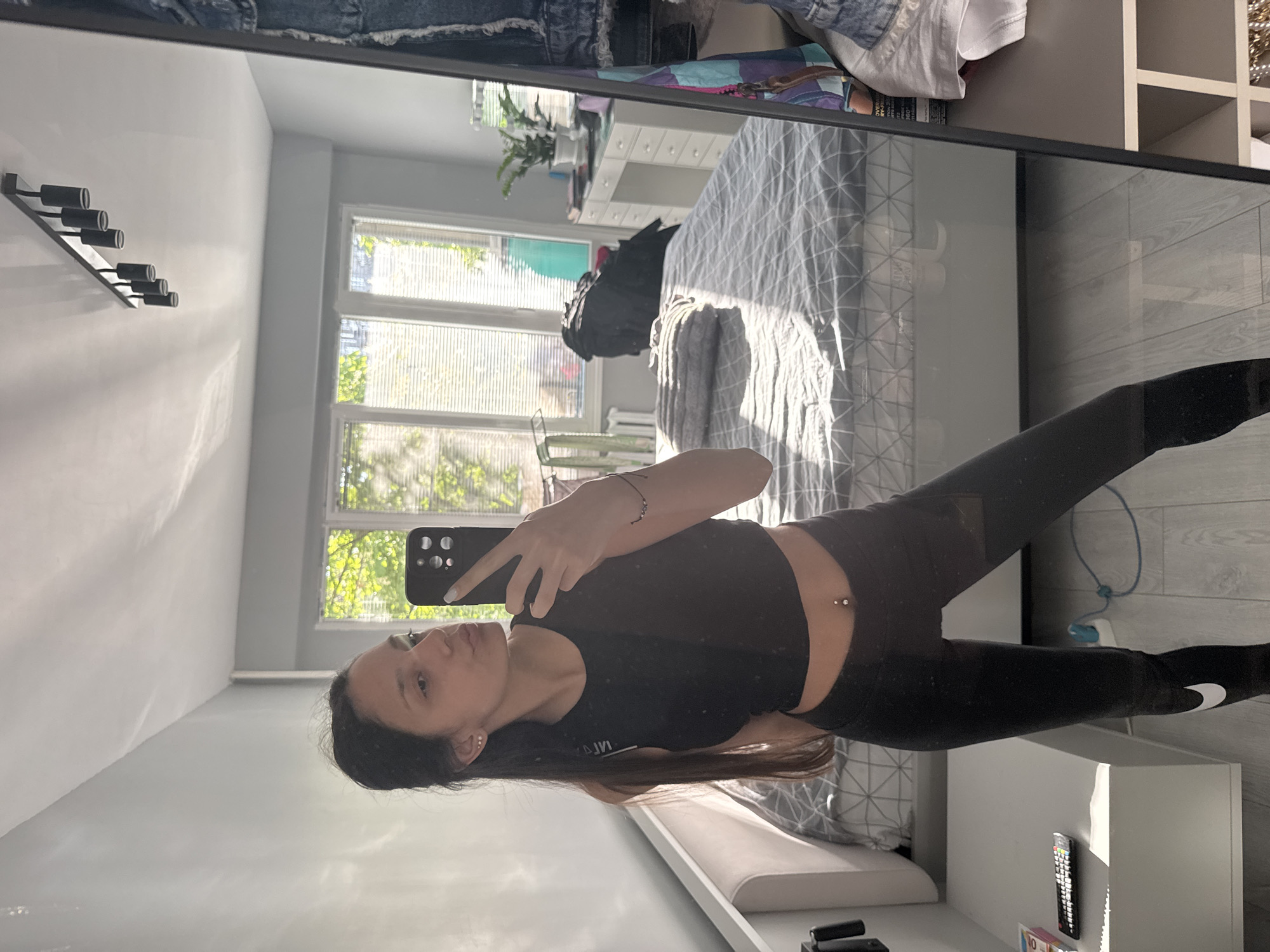 A young woman is taking a selfie in a mirror. She is wearing a black crop top and black leggings with a Nike logo on the ankle. Her long dark hair is partially visible. The background shows a bedroom with a bed, a window with blinds, a dresser, and a wall-mounted light fixture. While it is impossible to determine the location in the city of Sofia, Bulgaria from this image alone, the scene appears to be a modern apartment or home.