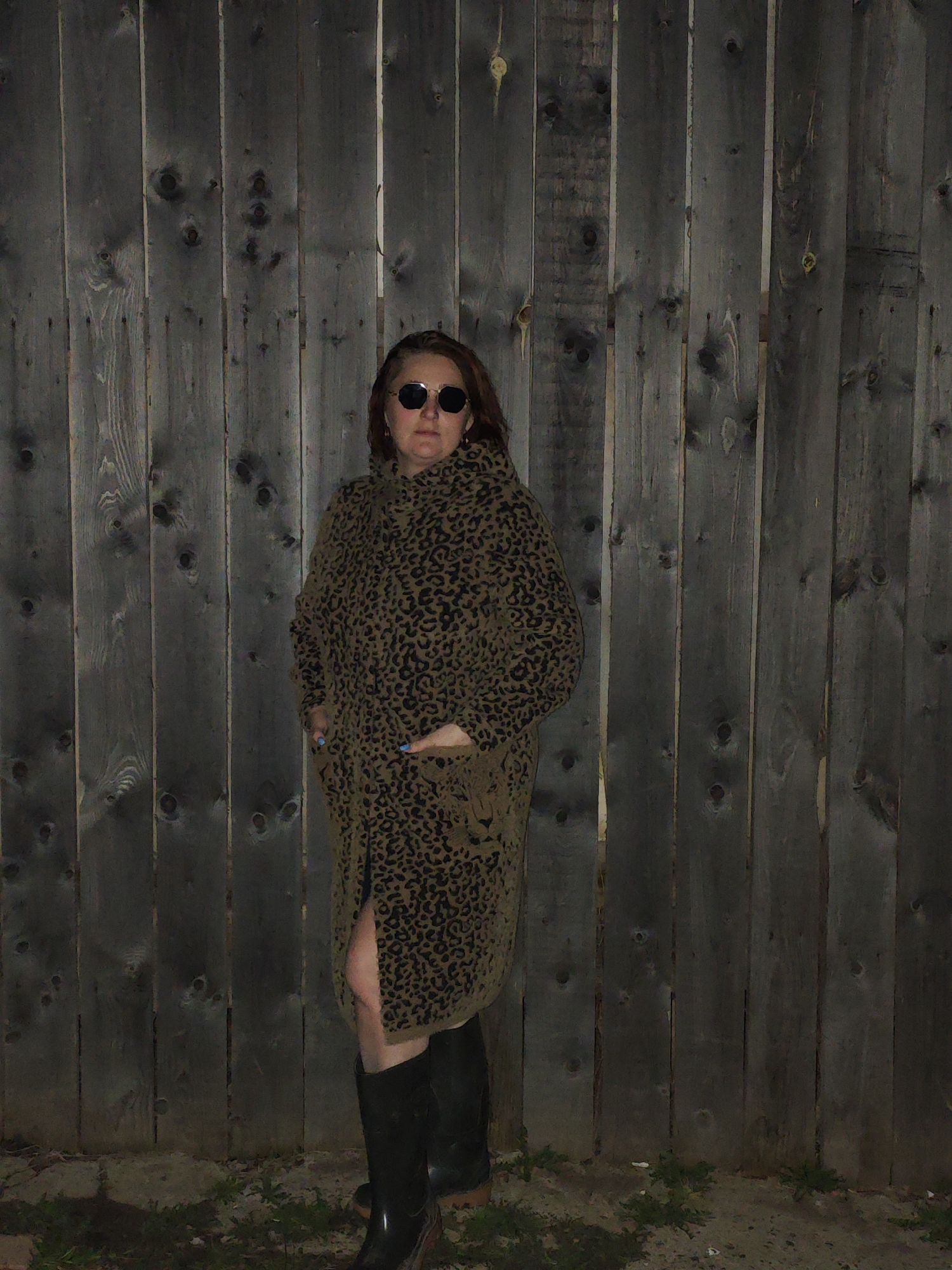 A person stands in front of a wooden fence. The person wears dark glasses, a long leopard-print coat, and tall black boots. The location appears to be outdoors at night. The ground is visible in the lower portion of the image and has some plant life. The fence is made of vertical wooden planks, and its weathered appearance suggests it is an older structure. The image is low-lit, and the details are somewhat obscured by the darkness. It's not possible to verify location in Фоки, Russia.