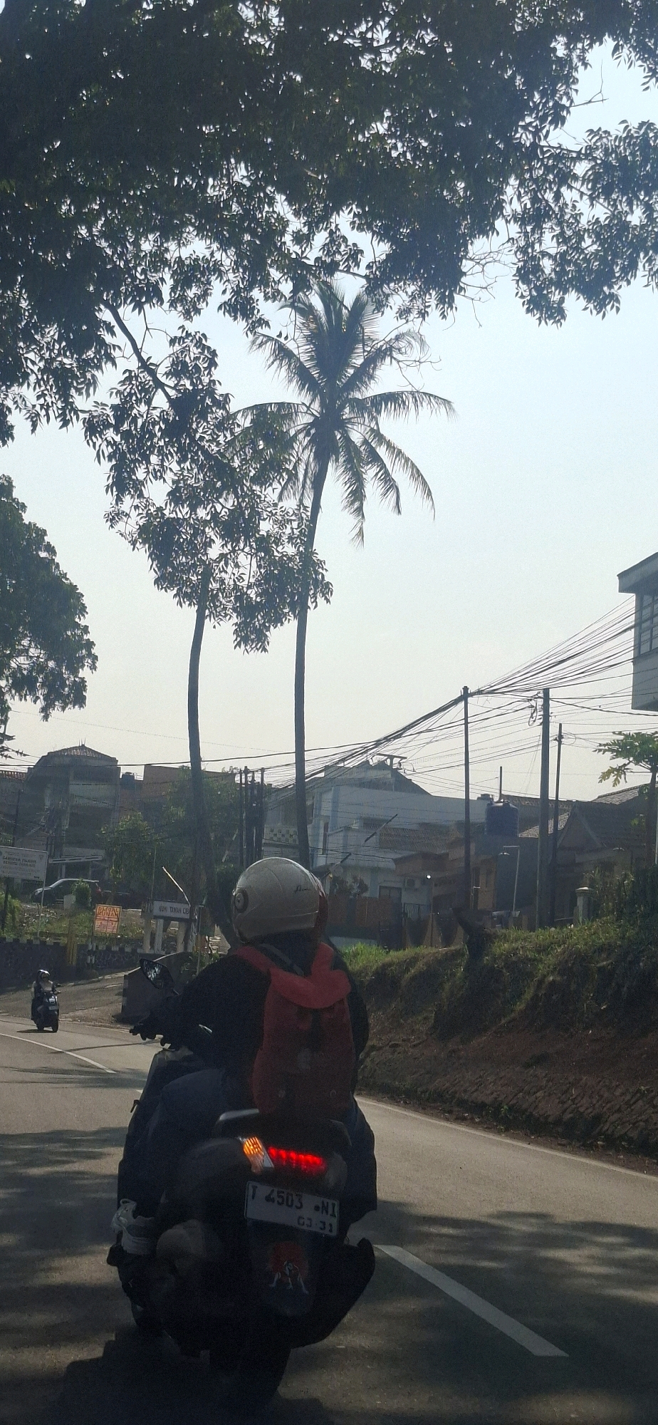 In the city of Cilaku, Indonesia, a person wearing a white helmet and a red backpack is riding a motorcycle on a paved road. The motorcycle has a license plate that reads "T 4503 NI" and "03-31". The rider is facing away from the camera, and the motorcycle's brake lights are illuminated. In the background, there are trees, buildings, and utility poles with numerous wires. Another motorcycle is visible in the distance, further down the road. The sky is clear and bright, suggesting it is daytime.