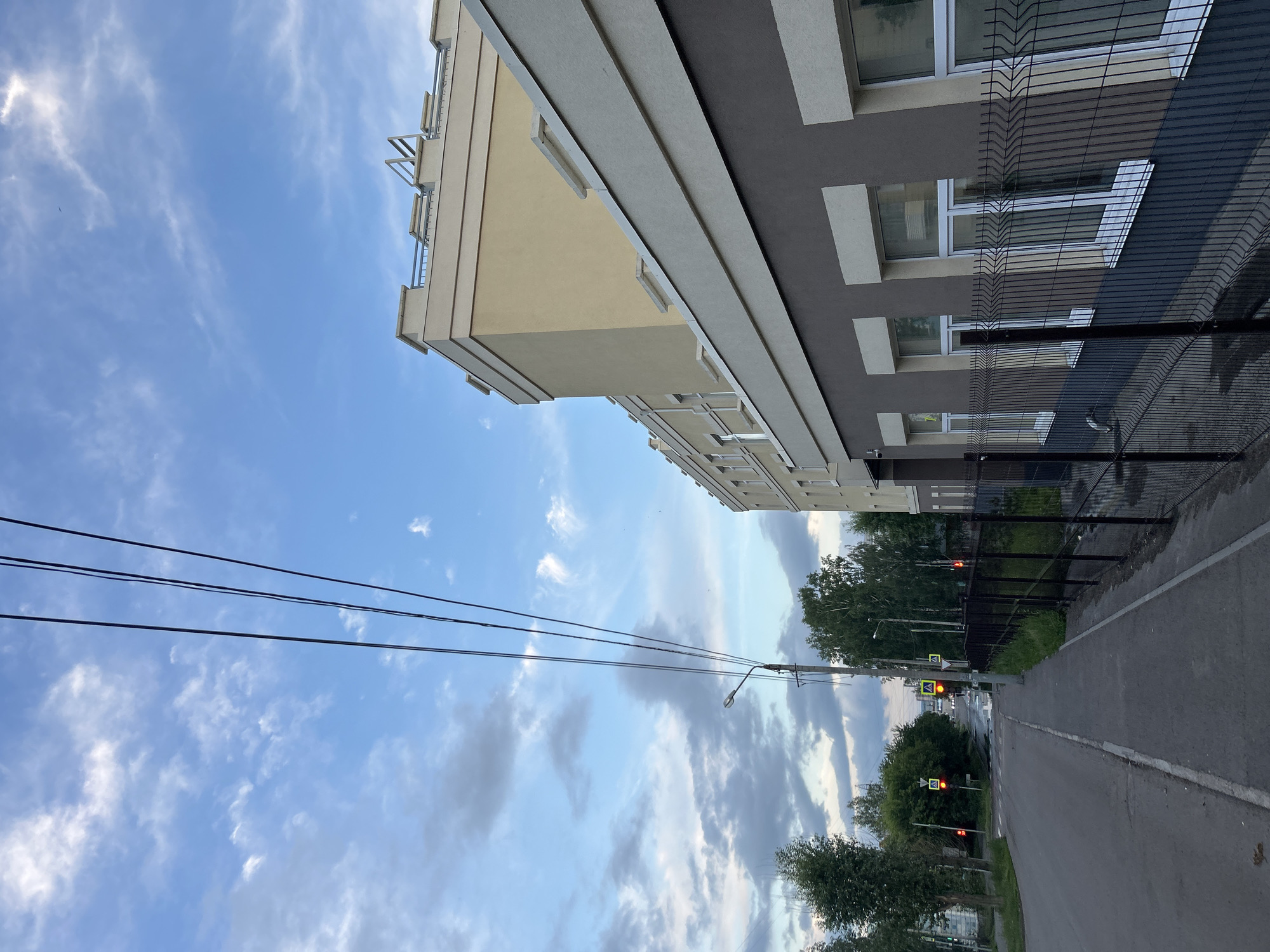 This media file displays a vertical, low-angle shot of a modern apartment building with a light yellow and dark grey facade. The building features numerous windows with white frames and a black metal fence runs along its side. To the left of the building, a clear blue sky with scattered white clouds fills the frame, with power lines crisscrossing the upper portion.
In the background, a paved road curves to the right, flanked by lush green trees. A street light and traffic signals are visible along the road. One traffic signal displays red lights, indicating a stop. The scene appears to be set during the daytime, possibly late afternoon or early evening, given the lighting and cloud cover. No people are visible in the frame. The overall impression is one of an urban environment with a contemporary residential building and surrounding greenery.
