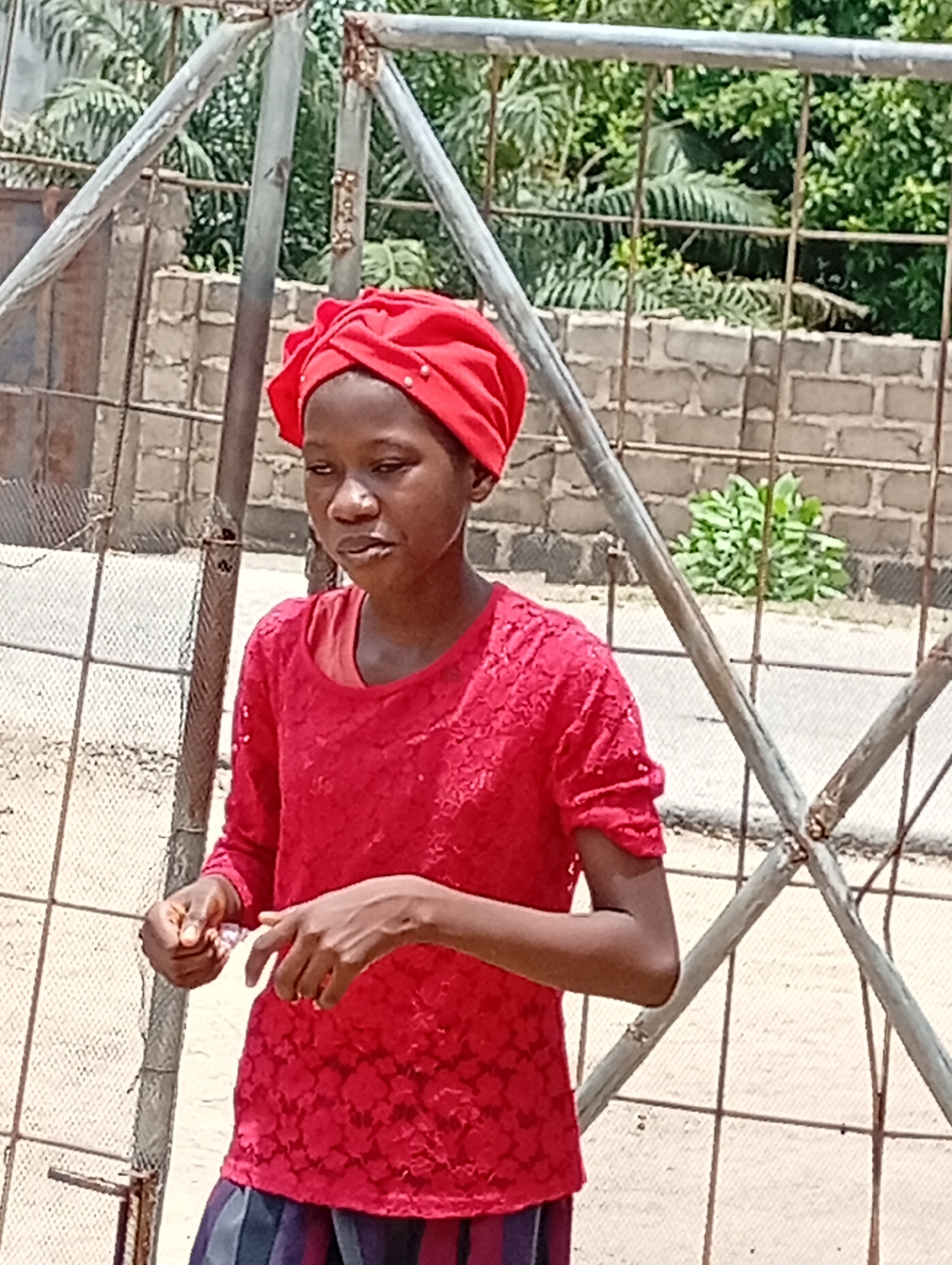 A young person with dark skin is standing behind a dilapidated metal fence in what appears to be an urban environment in Lokoja, Nigeria. The person is wearing a bright red headwrap and a matching red, lace-patterned top, with a glimpse of a striped garment visible at the bottom. Their gaze is directed slightly to the left, and their hands are occupied with a small object. The fence is constructed of metal bars and appears weathered, with visible rust and corrosion. Behind the fence, there is a brick wall and green foliage, suggesting a residential or semi-urban area. The background includes a paved road and additional greenery in the distance. The lighting is bright and appears to be natural daylight.