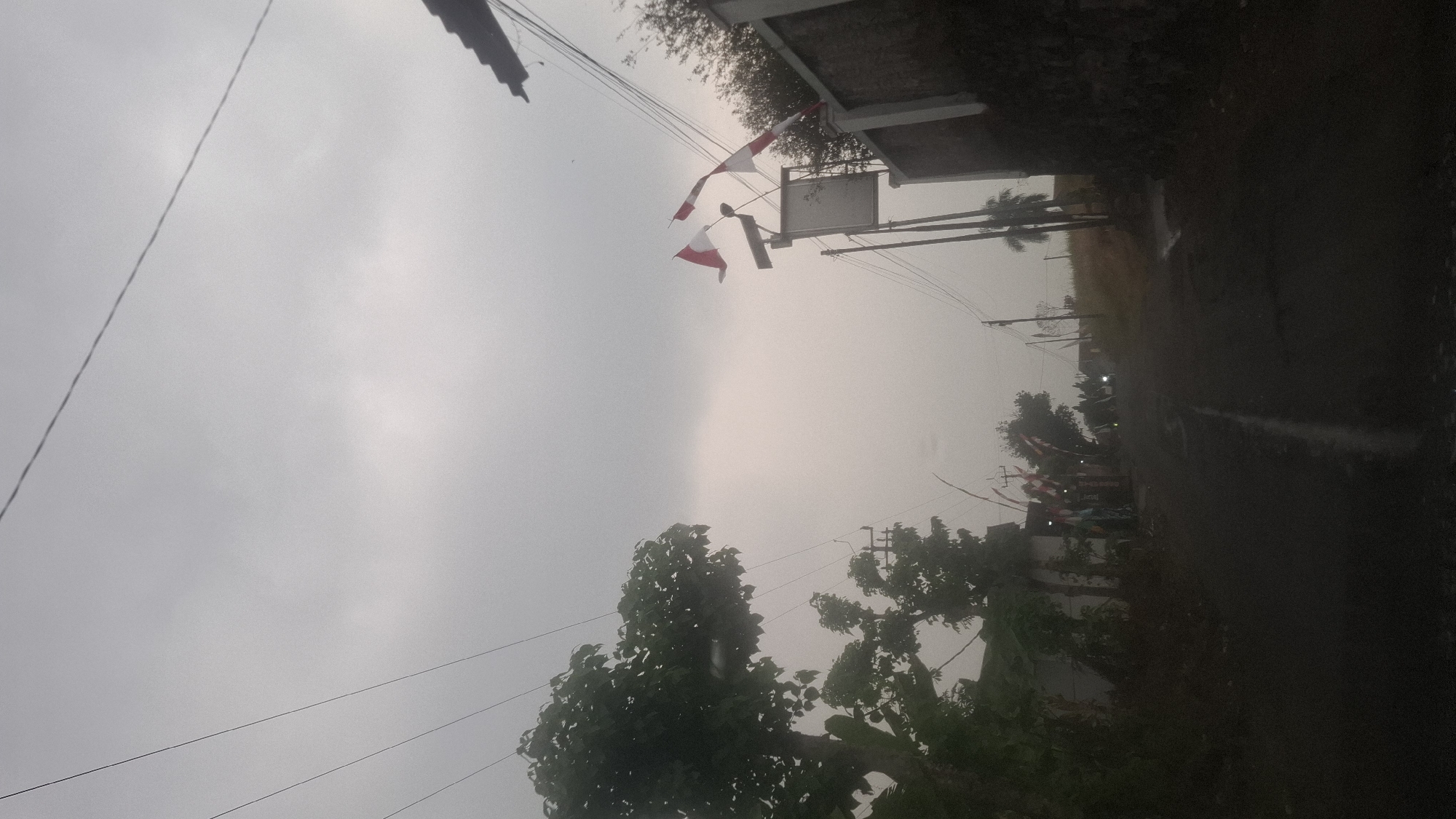 The image shows a low-angle, slightly underexposed view of a street scene in Cibinong Hilir, Indonesia, under an overcast sky. The road, which appears wet, slopes upwards into the distance. On the left side of the road, there are trees and houses. On the right side, a wall is visible, with a tall pole topped by two Indonesian flags flying. Further down the street, more flags are seen, and there are several motorcycles. The overall atmosphere is subdued due to the cloudy weather and low light.