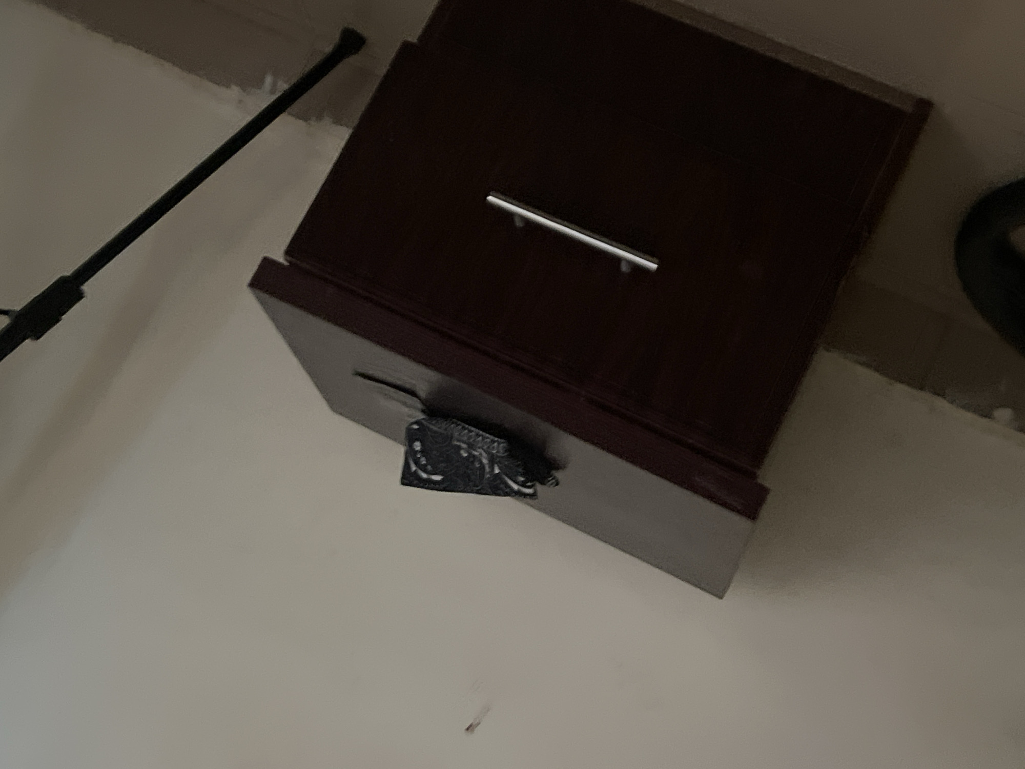 The image captures an indoor scene in what appears to be a room in Howayi, Nigeria, featuring a dark brown wooden cabinet or drawer unit positioned at an angle. The cabinet has a contemporary design with a single, slender, silver-colored horizontal handle on its front. Below the handle, a portion of the cabinet, possibly a drawer, is slightly ajar. Attached to the underside or edge of this partially opened section is a decorative black object, intricately patterned with white or silver designs that resemble floral or tribal motifs.
To the left of the cabinet, a long, slender black pole or rod extends diagonally across the frame, suggesting it could be part of a light stand, monopod, or another household item. In the upper right background, a dark, circular object is partially visible, possibly a wheel or the base of another piece of furniture. The room features light-colored walls, which appear somewhat textured. The lighting in the scene is dim, indicating either low artificial light or a time of day with limited natural light. No specific human activity is visible, and there is no discernible text in the image.