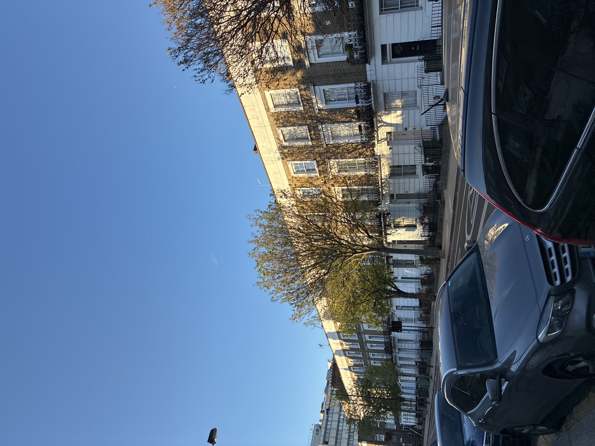The image depicts a street scene in London, United Kingdom, characterized by a row of townhouses with uniform architectural style, distinguished by brick and white facades. These buildings feature symmetrically arranged windows, wrought iron railings, and modest front entrances. A mature tree stands between the buildings, casting shadows across the facades. The sky is clear and blue.
Foreground elements include two parked vehicles, a dark SUV and a gray SUV. A partial view of another dark-colored car is visible. A street lamp is situated near the bottom-left corner. The overall scene is bathed in sunlight, suggesting a bright, daytime setting.