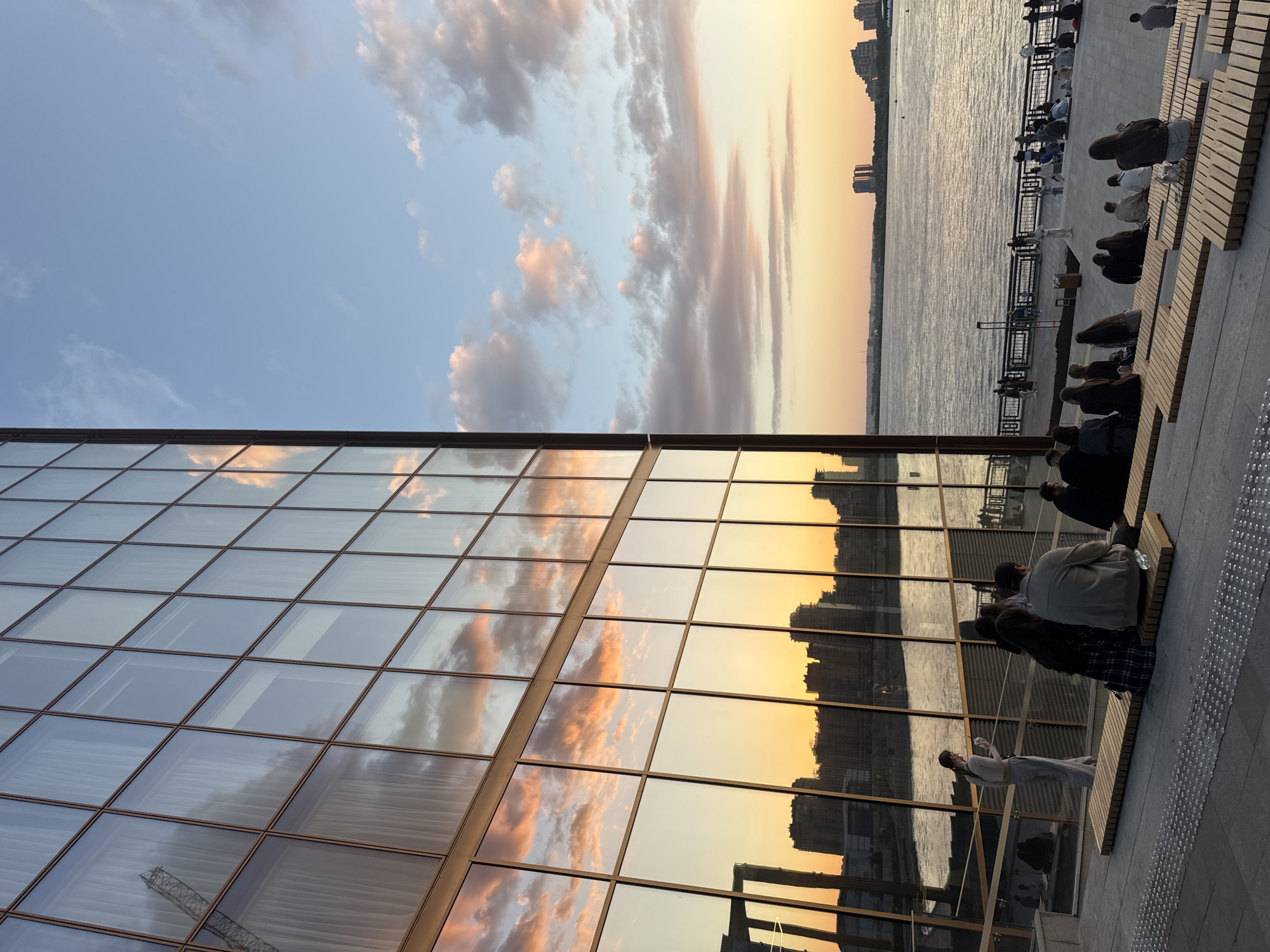 The image captures a panoramic view of a riverfront embankment at sunset, specifically located in Kazan, Russia, along the Kazanka River near its confluence with the Volga, in the vicinity of the Kazan Family Center and Rivera hotel complex.
Dominating the left side is a tall, multi-story building with a full glass facade, segmented by thin, dark brown frames into a grid pattern. The reflective surface prominently displays the vivid colors of the sunset sky, transitioning from a soft blue at the top with faint white clouds and a faint reflection of a construction crane, to vibrant orange and pink clouds in the middle, and a golden-orange hue reflecting the distant city skyline and the river at the lower levels. White curtains or blinds are faintly visible through some of the reflected glass panels.
To the right, the wide expanse of a river flows under a sky painted with the warm tones of dusk. The upper right sky is a clear light blue, gradually shifting to pale orange and pink near the horizon, dotted with scattered, elongated clouds that catch the sunset light. The river's surface is a shimmering dark expanse, reflecting the sky's colors and showing subtle ripples. The opposite bank features a low-lying, dark silhouette of a city, with numerous buildings and two distinct taller structures visible on the far right.
In the foreground and midground, a diverse group of people are gathered on a tiered public seating area constructed from light-colored wooden planks set into a darker stone or concrete foundation. Many individuals and small groups are seated, largely facing the river and the sunset. A woman in light-colored casual attire stands near the building, looking at a mobile phone. Further back, along a dark metal railing separating the seating area from the river, more people are visible, standing or sitting on benches, enjoying the evening atmosphere. The overall scene depicts a serene urban leisure space during twilight.