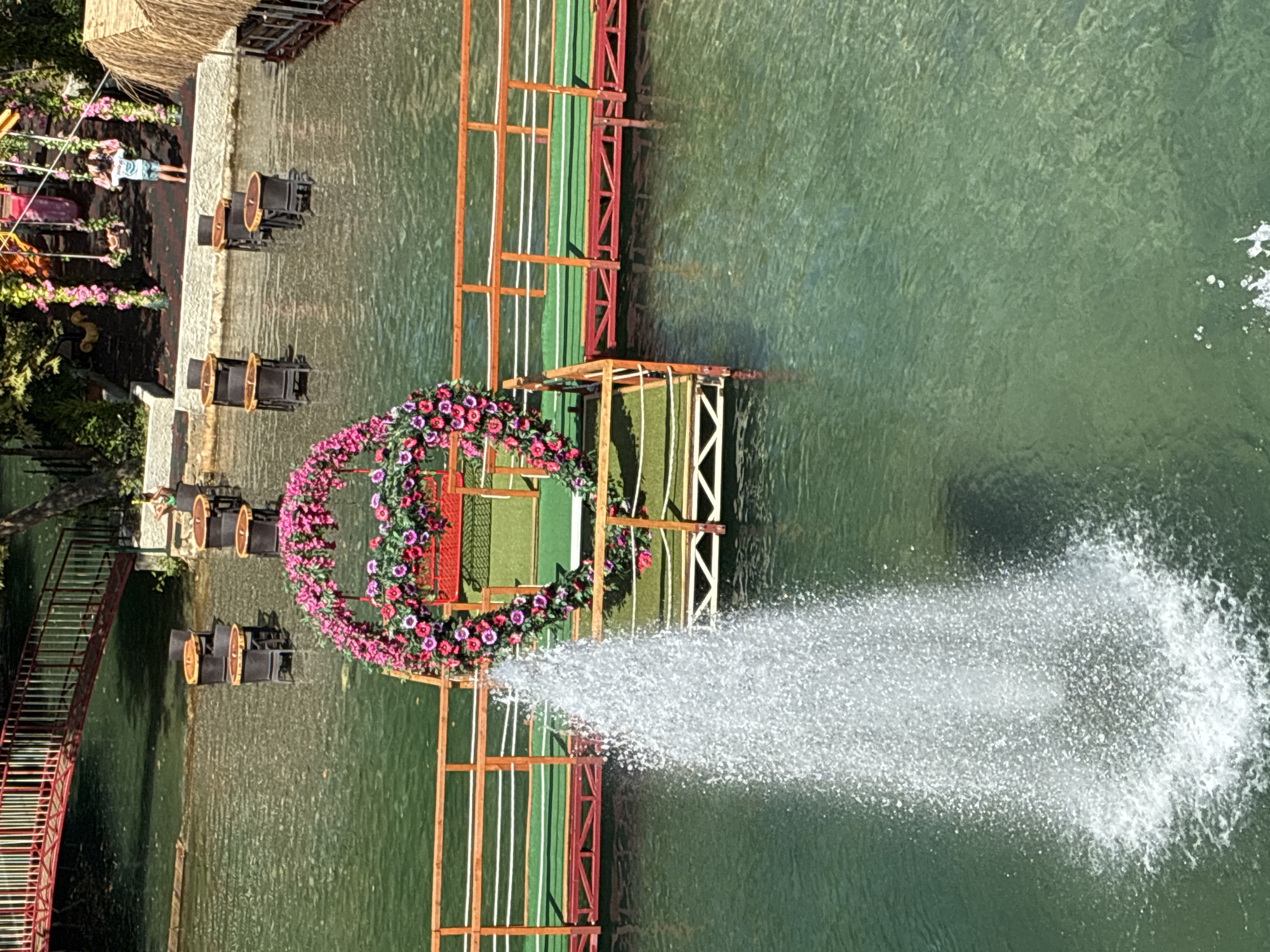 This image depicts a serene outdoor dining area situated on a body of water, likely in Alanya, Turkey. In the foreground, a fountain erupts from the clear green water, its spray creating a misty curtain. A floral archway, adorned with vibrant pink and red flowers, frames a section of the water.
The area features wooden platforms and walkways with red railings and green carpeting, suggesting a dedicated recreational or dining space. Several tables and chairs are submerged in the shallow water, creating a unique dining experience. A red bridge with a white railing extends over the water in the upper left corner, hinting at further connections or viewing points.
In the background, beyond the submerged tables, a thatched-roof structure is visible, possibly a restaurant or bar. Further back, a playground with swings and colorful structures is apparent, with a young person on one of the swings. The presence of lush green trees and vegetation indicates a natural and scenic setting. The water itself appears calm and inviting, reflecting the surrounding greenery and structures.