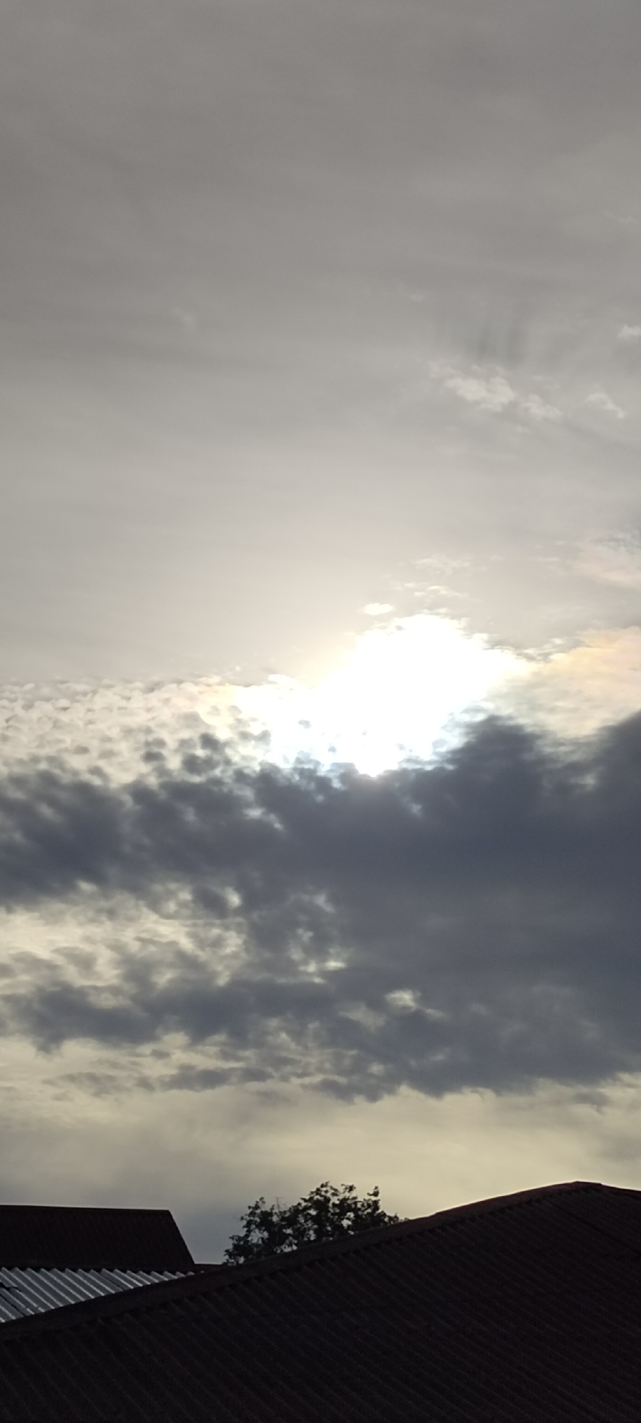 The image shows a sky partially obscured by clouds, with a bright area suggesting the sun behind the cloud cover. Below the clouds, the roofs of buildings are visible, including corrugated metal and darker tiled surfaces. A small tree is positioned between the roofs. The location is Meunasahgeumata, Indonesia. The scene depicts a cloudy day in an urban environment. There are no people visible in the image.