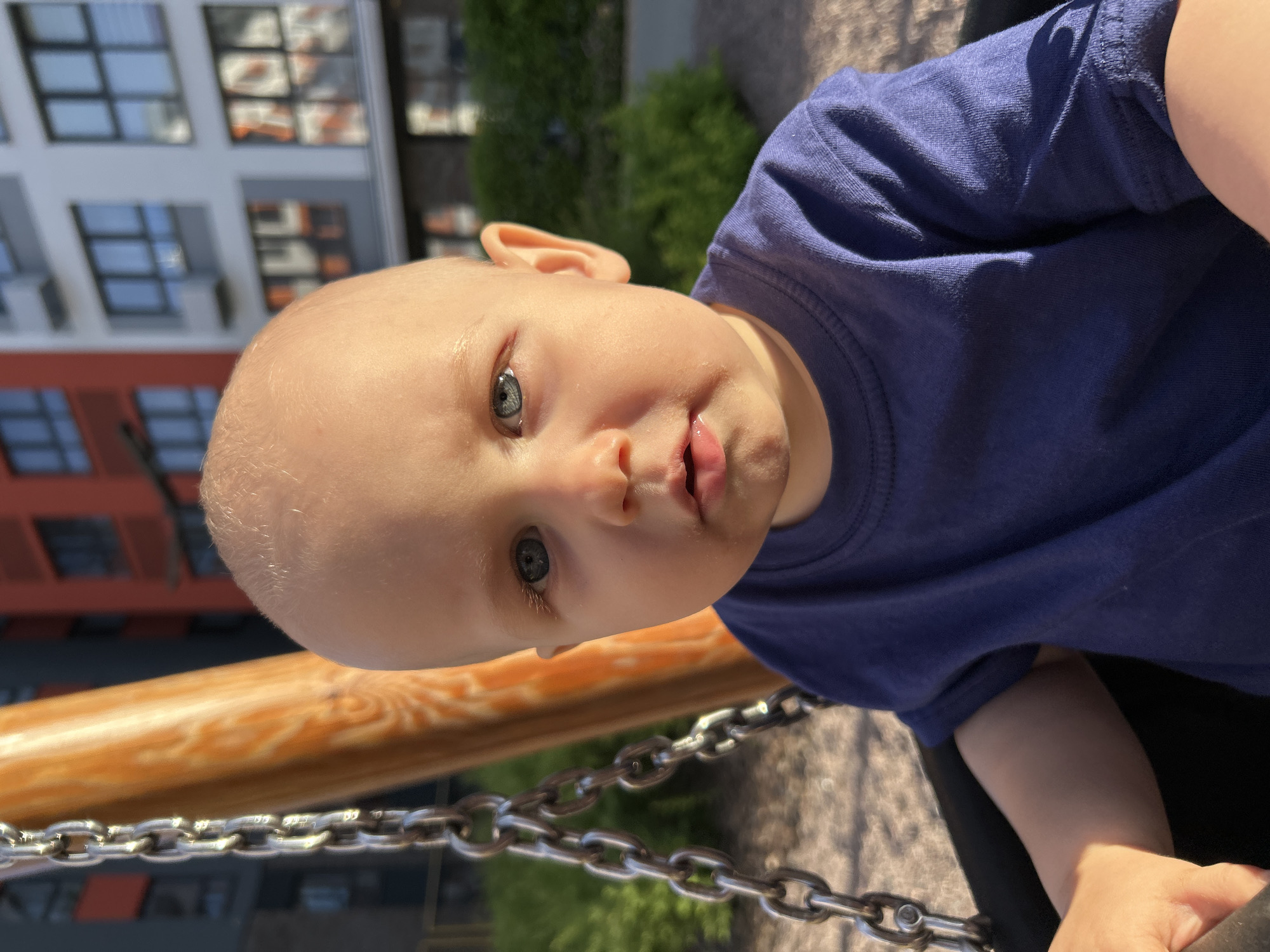 A close-up, vertically oriented shot features a baby with light skin and blue eyes. The baby is wearing a dark blue t-shirt and is looking upwards towards the camera. The baby's head is tilted, and their mouth is slightly open. In the foreground, a wooden bar is visible, with a metal chain running beneath it. The background is blurred, but suggests an outdoor setting with a building that has many windows and green foliage. The lighting suggests it is daytime, and the weather appears to be clear. The image appears to be taken at a playground, possibly in Kaluga, Russia. There is no visible text in the image.