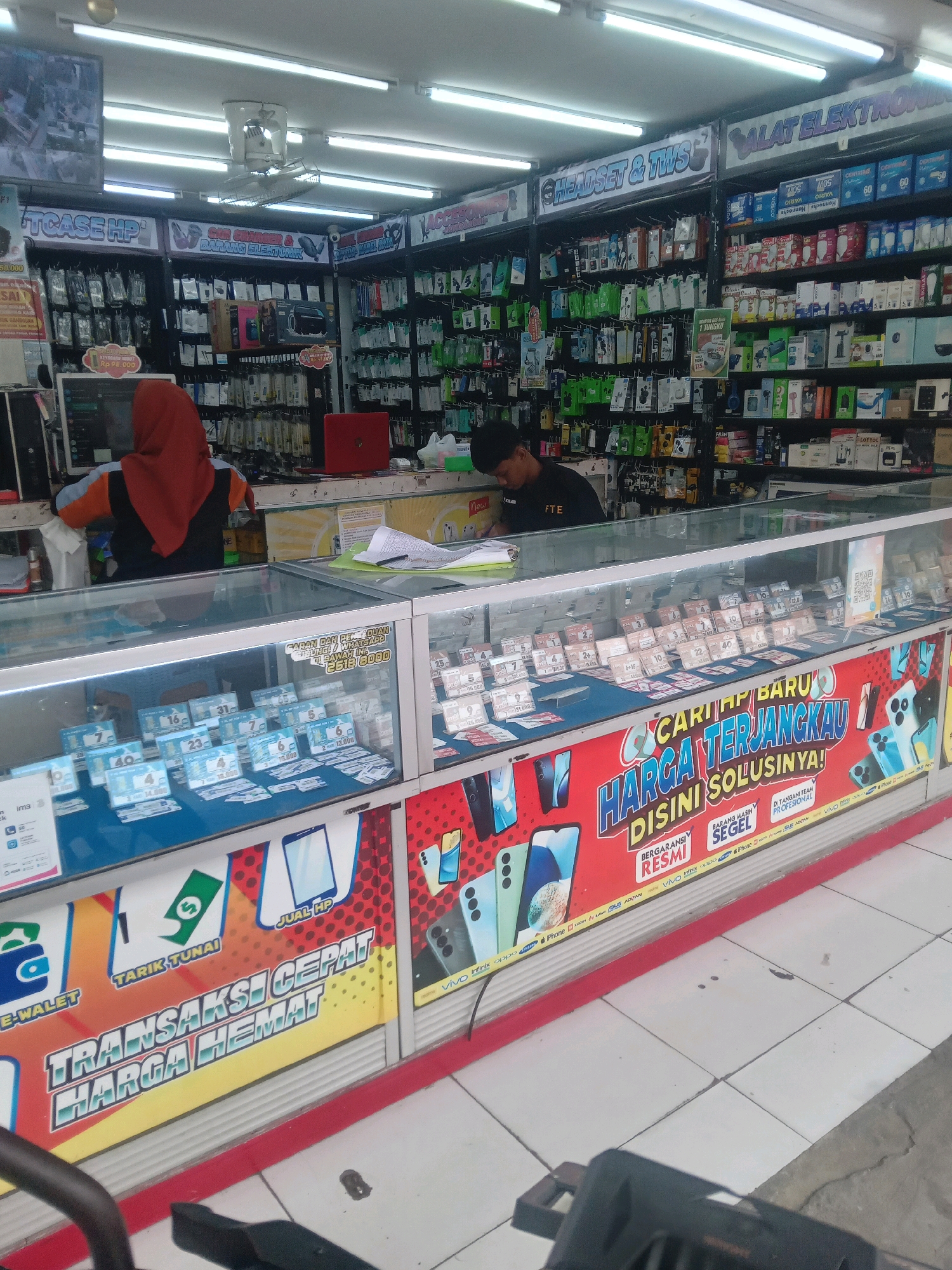 The image shows the interior of a brightly lit electronics store located in Depok, Indonesia. The store is packed with shelves displaying various electronic accessories, including phone cases, chargers, headphones, and other small gadgets. In the foreground, a glass display counter showcases a variety of prepaid phone credit cards and SIM cards. A person wearing an orange and black uniform, likely an employee, is visible behind the counter, interacting with paperwork. Another employee with dark hair is seated at a workstation on the left side of the frame, also engaged with documents. Prominent signage in the store features advertisements for mobile phones and electronic services, with some text in Indonesian. The flooring is tiled, and the overall impression is of a busy retail environment focused on mobile accessories and telecommunications services.