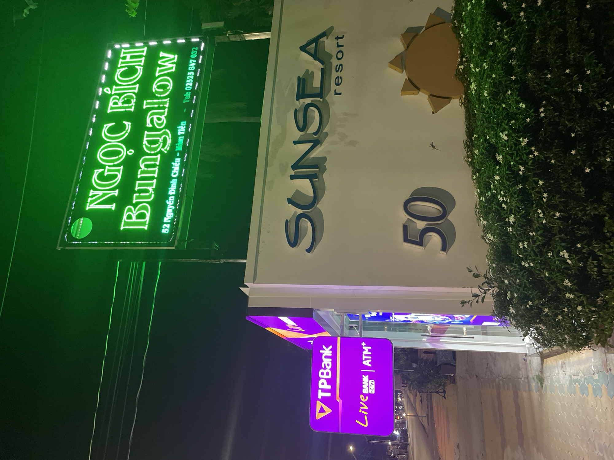 The image depicts a night scene in the city of Phu Thuy Ward, Vietnam, showcasing the illuminated signage of "Ngọc Bích Bungalow" and "SUNSEA Resort 50", along with a "TPBank ATM+" sign. The Ngọc Bích Bungalow sign is a rectangular board lit in green with text in Vietnamese. Adjacent to it is the SUNSEA Resort sign in silver with a golden sun logo. A purple TPBank ATM+ sign is present. The background includes streetlights, vegetation, and building facades, indicating an urban environment.